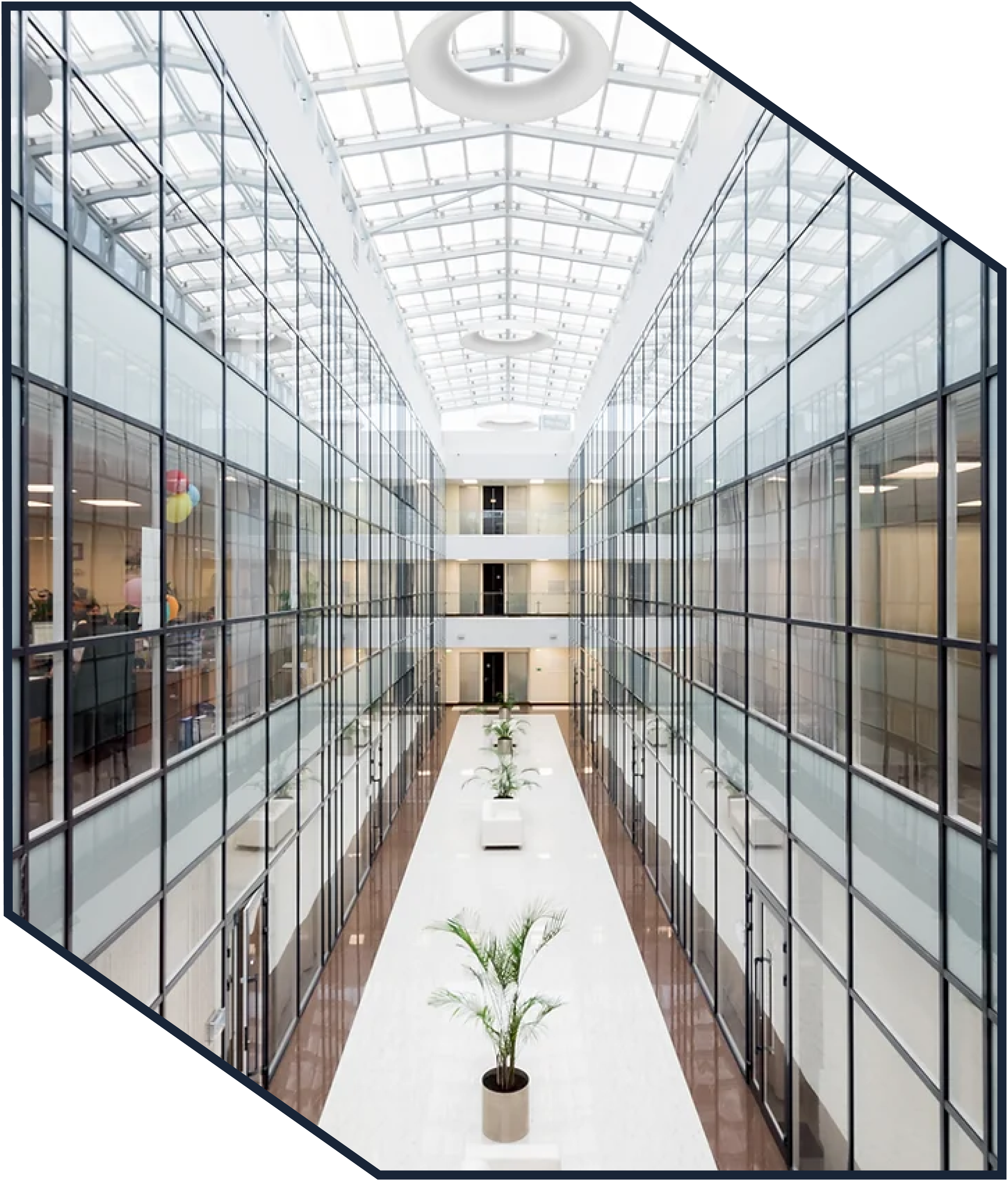 Interior view of a modern office building with glass walls, a high glass ceiling, and a central corridor with potted plants.