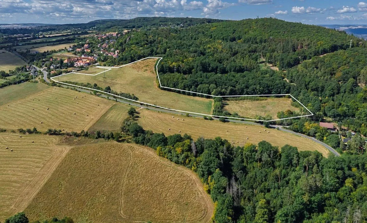 Aerial view of a rural landscape with a large wooded hill on the right, a white boundary outline around a section of flat farmland, roads, and a small residential area in the distance.