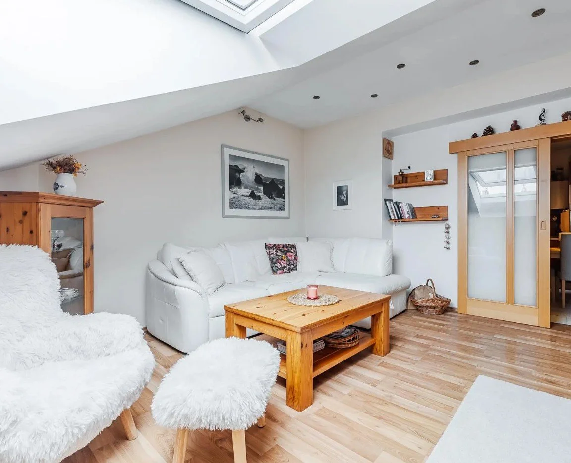 Living room with white sofa, wooden coffee table, fluffy white chairs, photogrpah on the wall, wooden display cabinet, and sliding door leading to another room under a sloped ceiling with skylights.