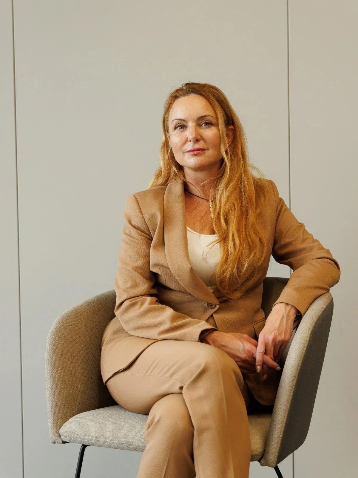 A woman with long red hair, dressed in a tan suit, sitting in a beige chair in front of a plain light wall.