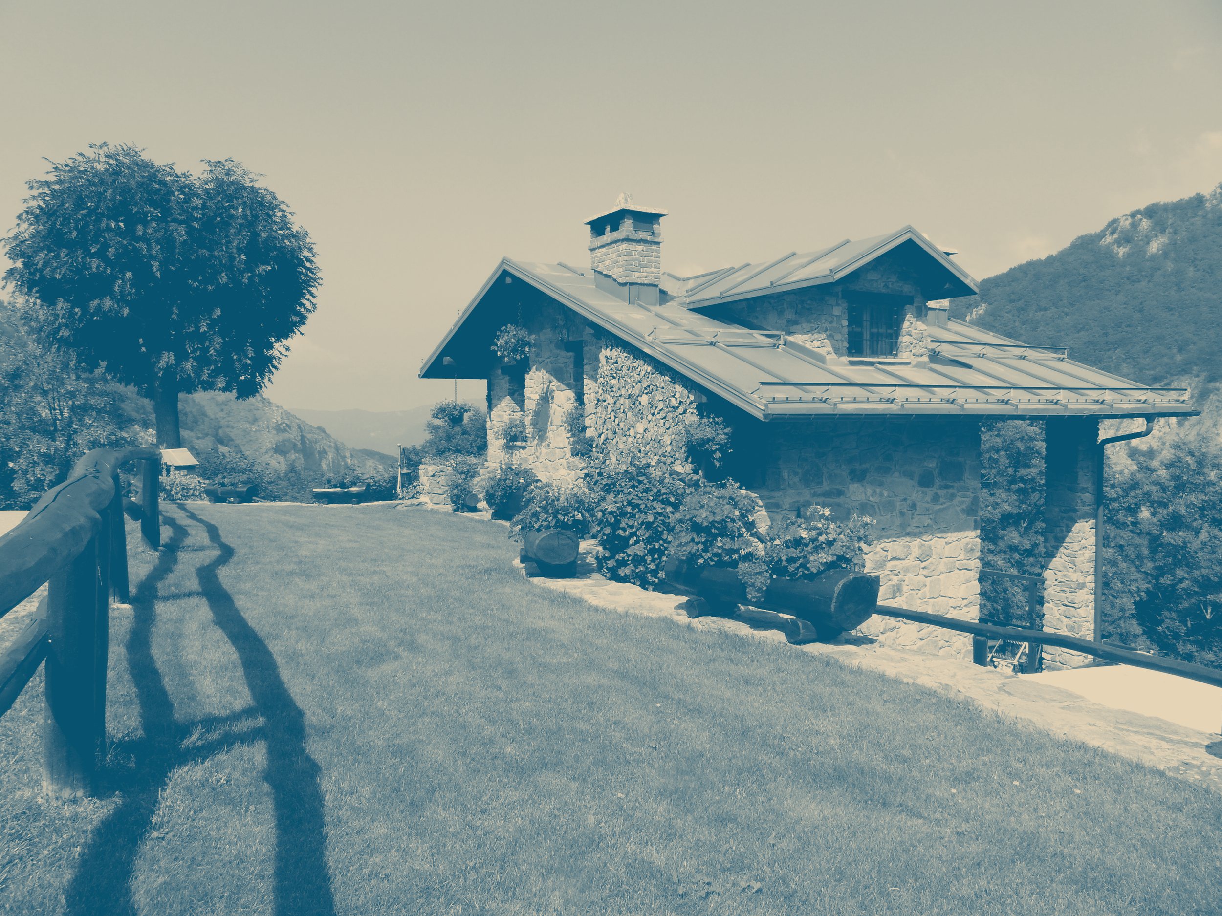 A stone house with a metal roof in a rural landscape, surrounded by trees and mountains, with a grassy path and wooden fence.