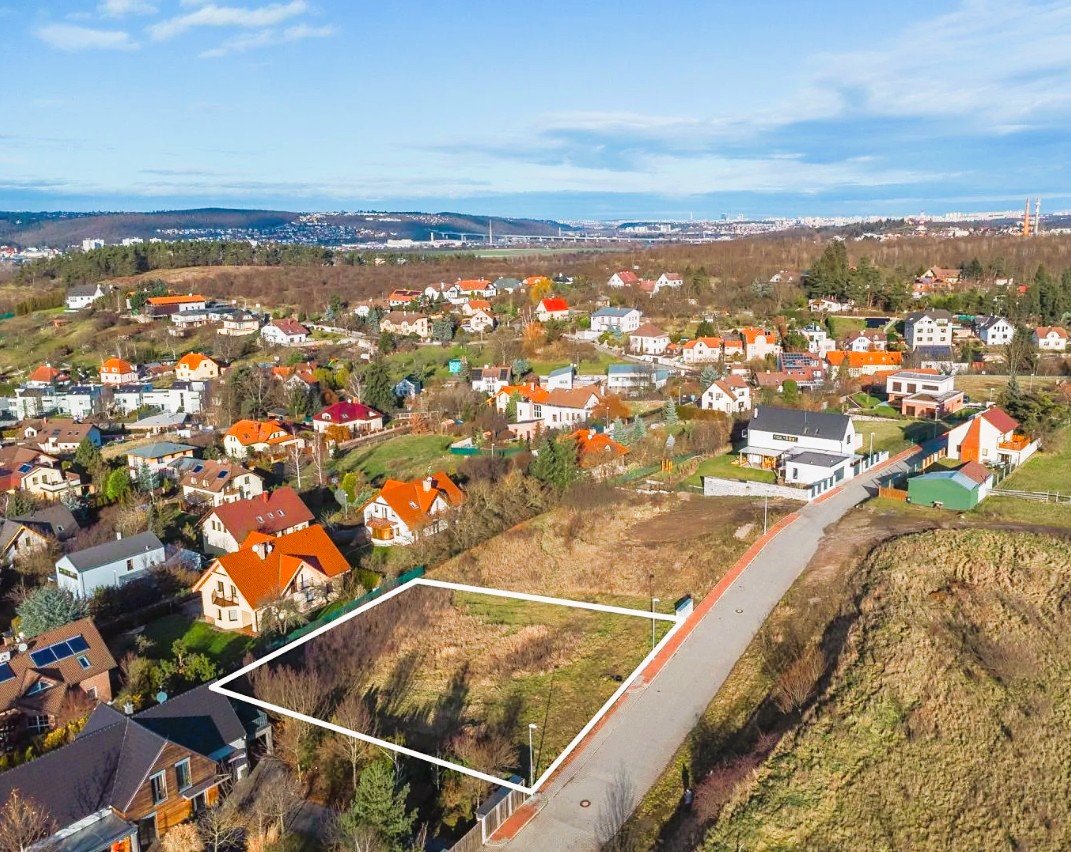 An aerial view of a residential neighborhood with houses, roads, and a vacant lot outlined in white. The neighborhood is on a hilly terrain with a mix of trees and buildings, with a city skyline in the distance under a partly cloudy sky.