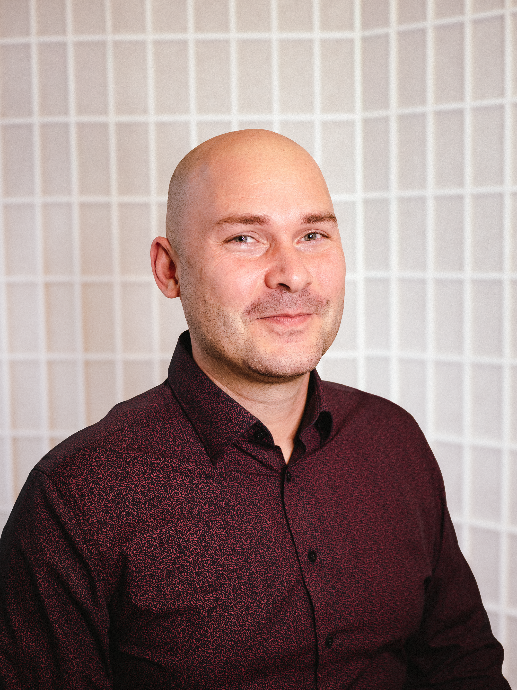 Portrait of a bald man wearing a dark patterned shirt in front of a grid-patterned background.