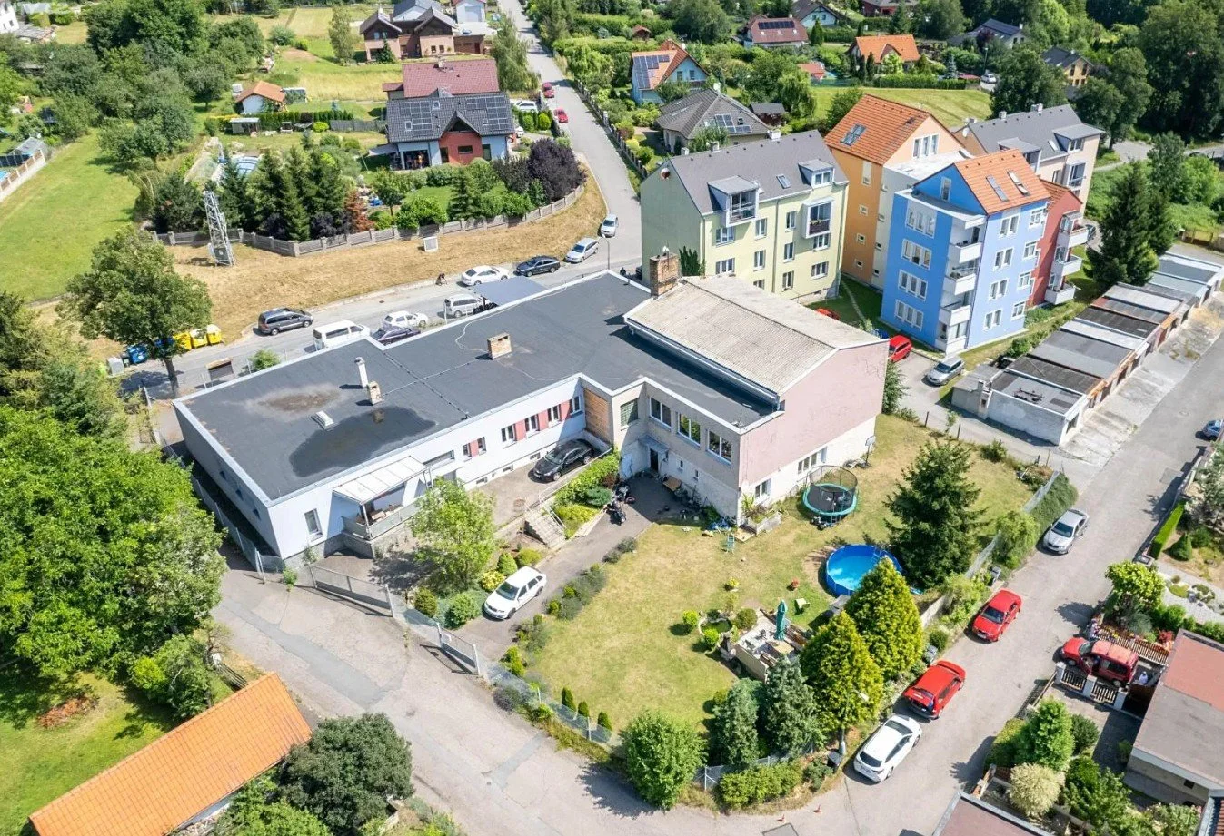 Aerial view of a residential neighborhood with houses, apartment buildings, parked cars, trees, and a grassy yard with a trampoline and a small pool.