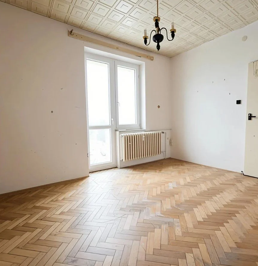 Empty room with white walls, a window, radiator, parquet flooring, and a black chandelier ceiling light.