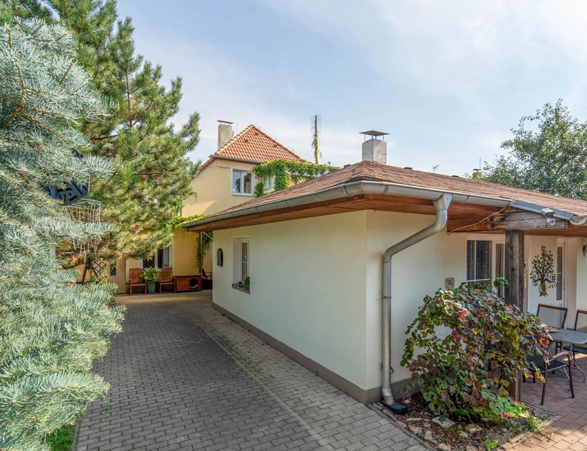 A house with a beige exterior, red-tiled roof, and a small porch area with outdoor chairs. There are trees and plants around the house, and a paved walkway in front.