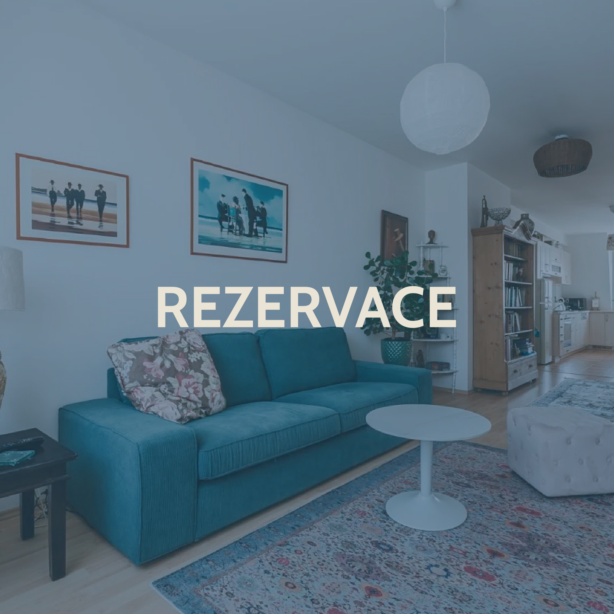 Living room with a blue sofa, floral pillow, coffee table, patterned rug, bookshelf, kitchen in the background, and the word 'REZERVACE' overlayed.