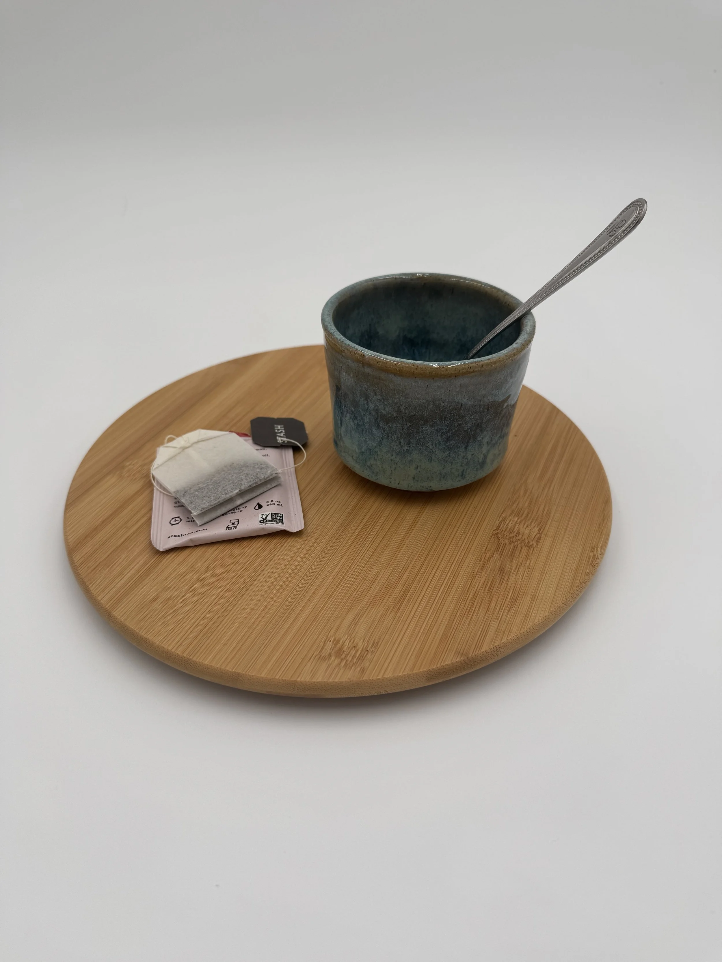 Handmade ceramic mug in cool blue and green Rainforest Storm glaze, sitting on a bamboo base with a teabag beside it and a spoon resting inside, waiting for a hot cup of tea.