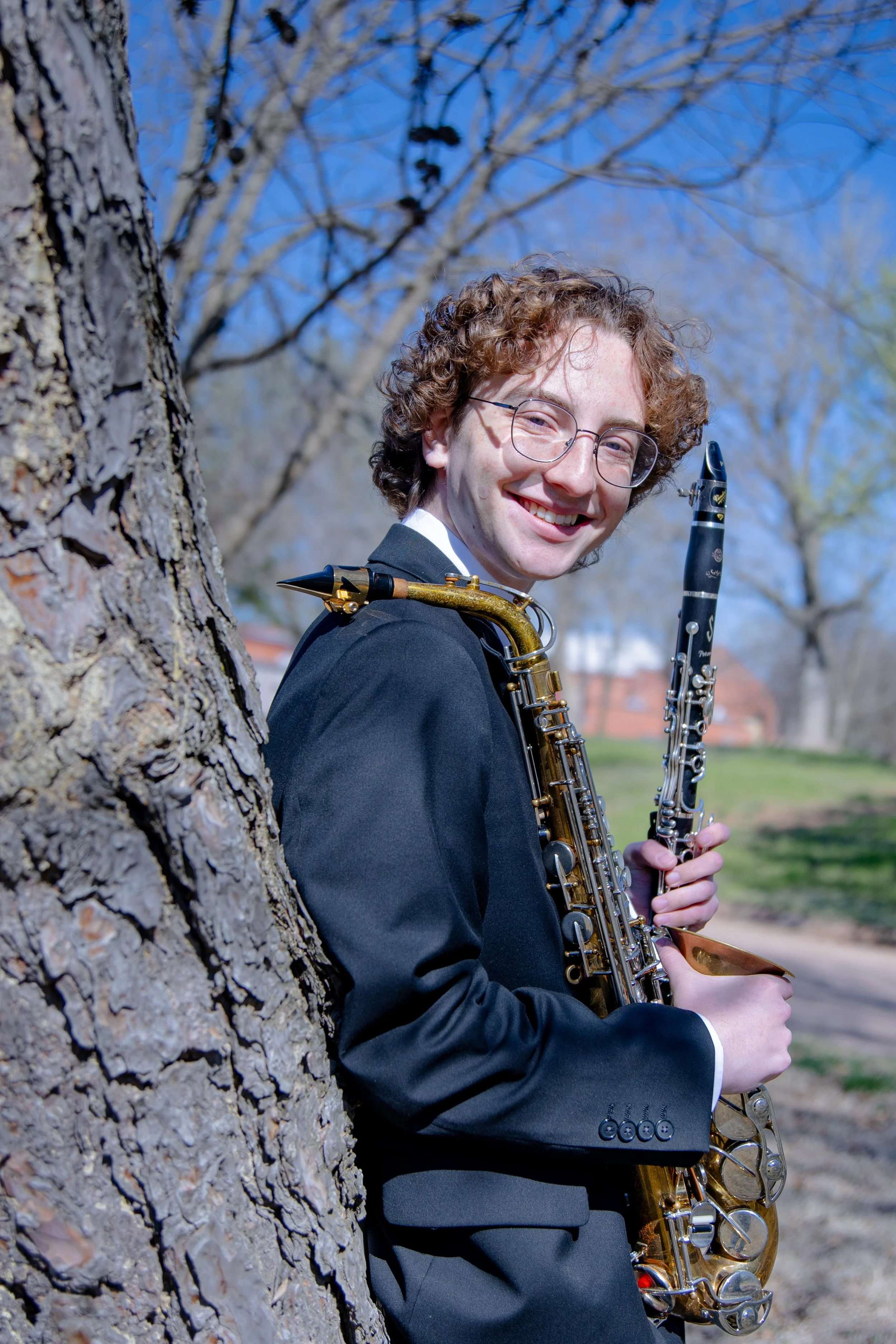 Kenidy Hutchins leaning against a tree on the Oklahoma Baptist University campus, holding a saxophone in his right hand and a clarinet in his left, linking to his portfolio featuring performances from his Sophomore Recital