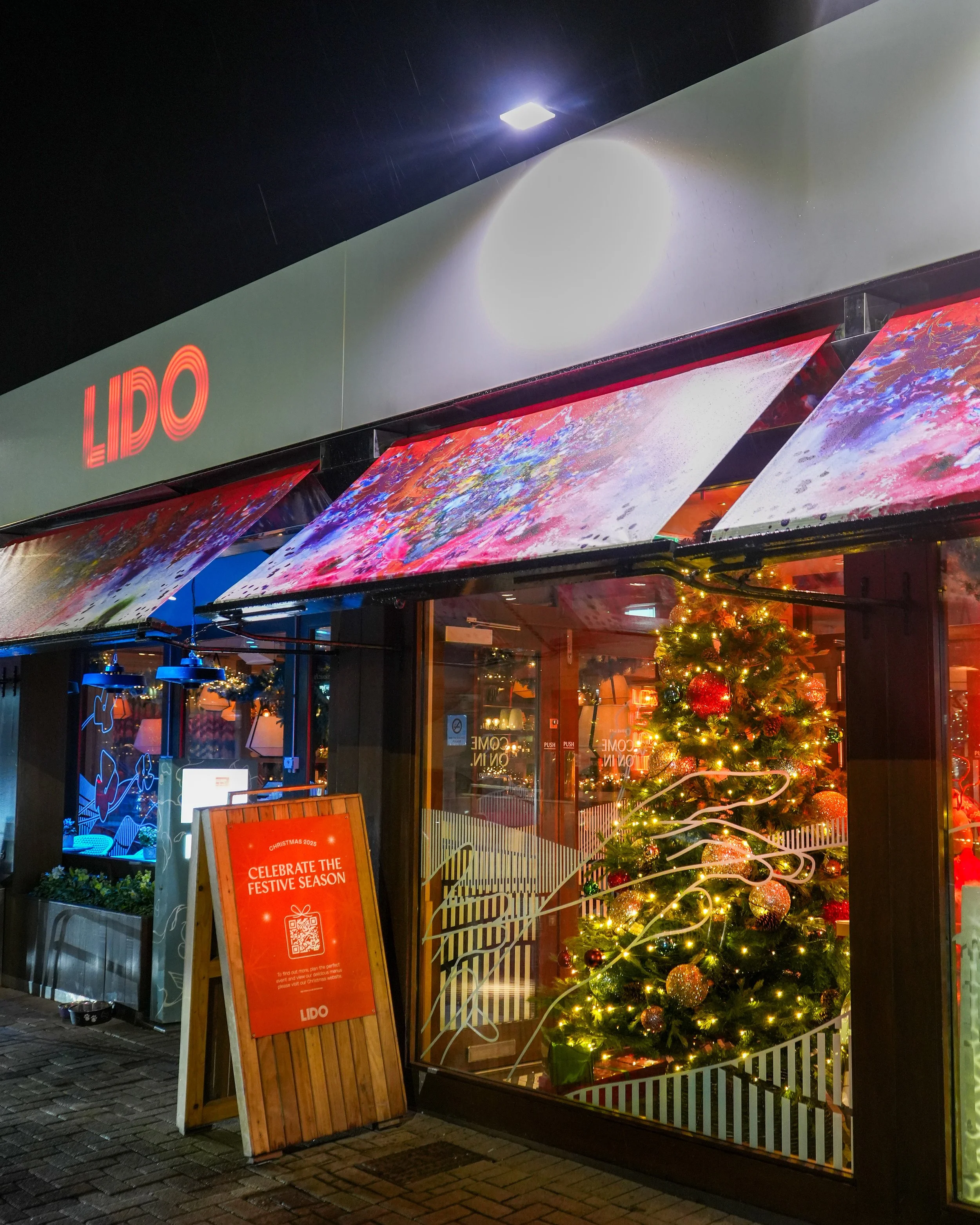 A Christmas decorated storefront with a Christmas tree inside, illuminated with decorations and lights. A red signboard outside reads, 'Celebrate the Festive Season' and includes a QR code. The store has a sign 'LIDO' in red on top.