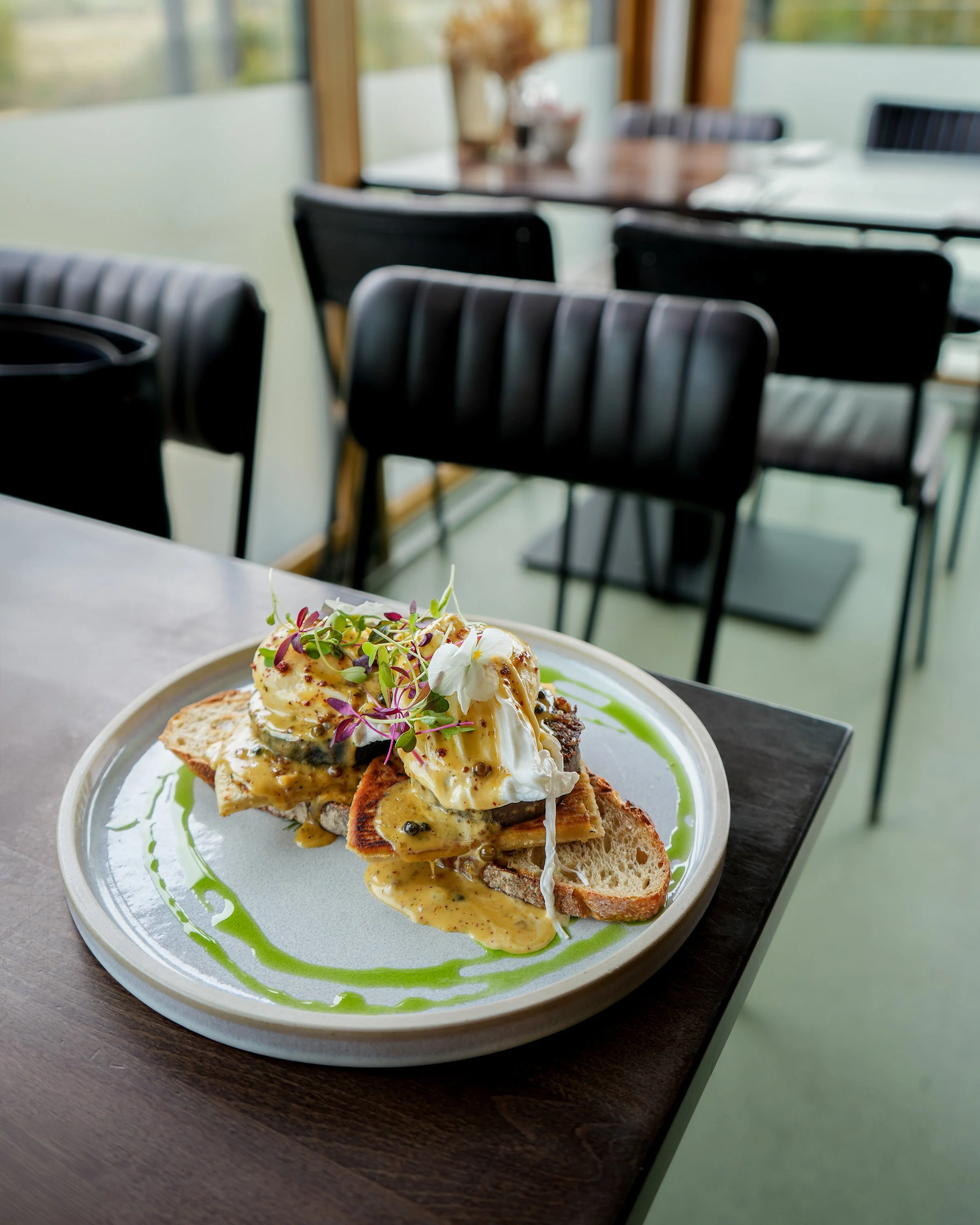 Plate of eggs Benedict with microgreens, green sauce, and bread slices on a table in a bright restaurant or café.