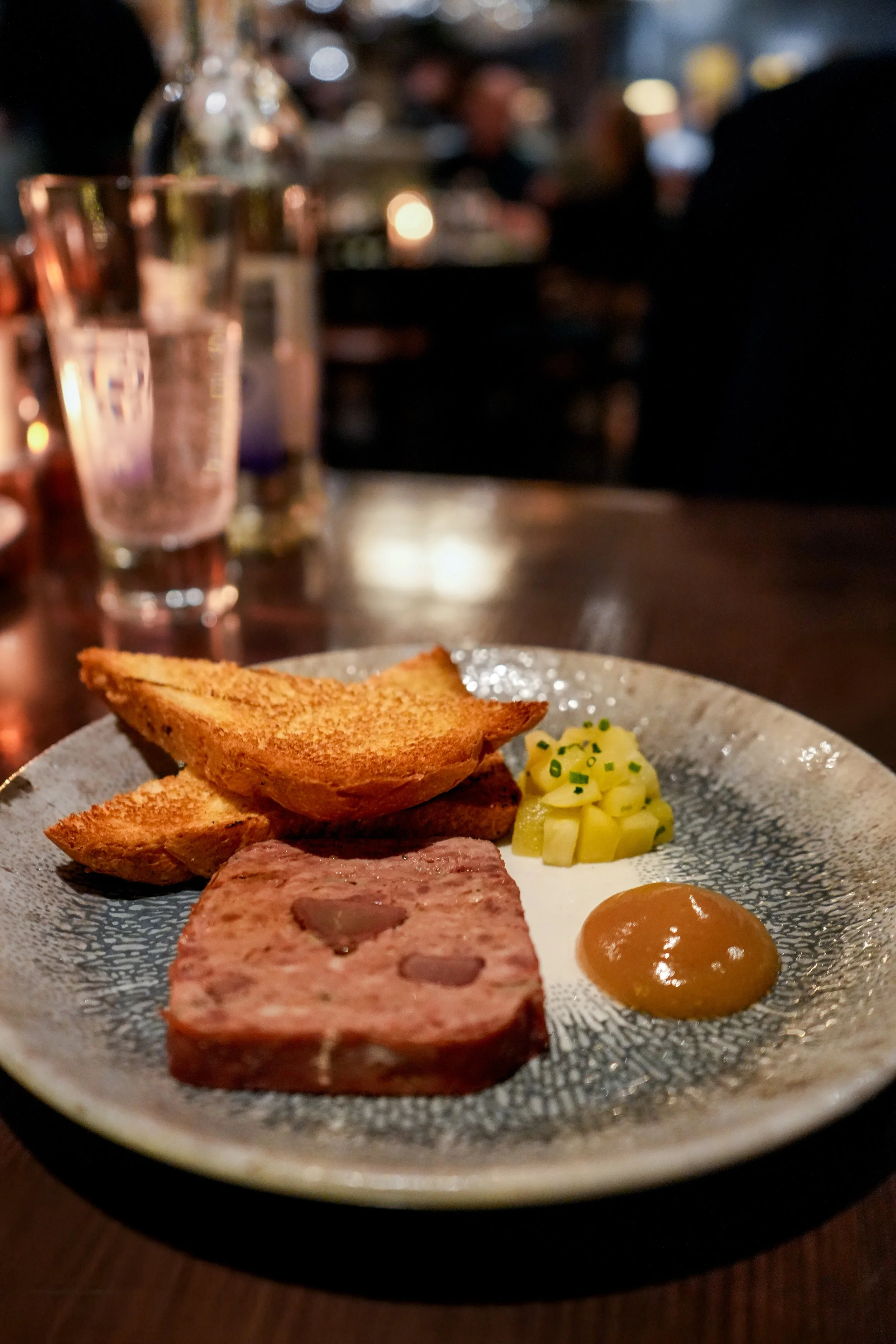 Plate with toast, pâté, pickles, and mustard on a table in a dimly lit restaurant.