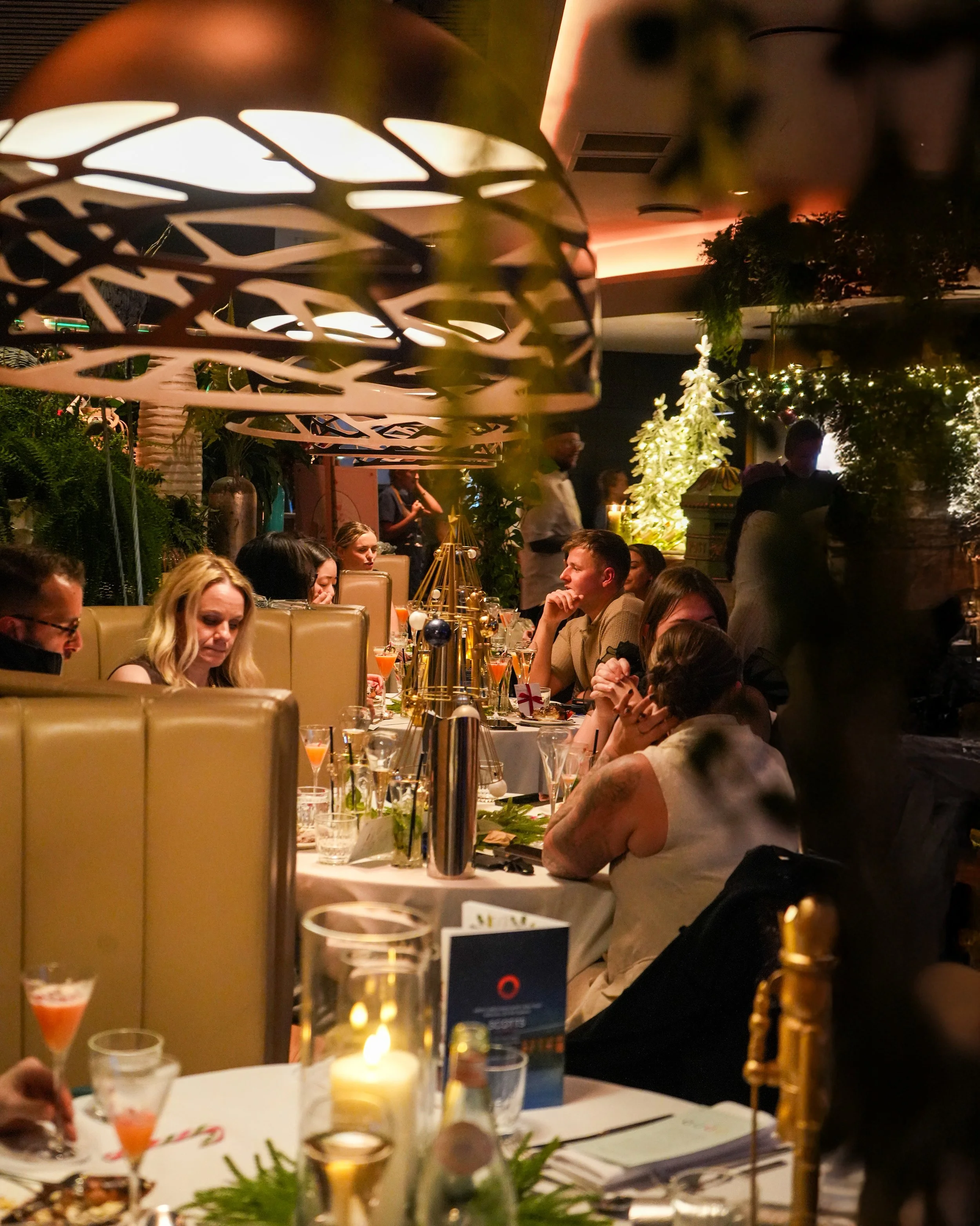 People sitting at a warmly lit restaurant table decorated with candles, drinks, and holiday decorations, including a Christmas tree in the background.