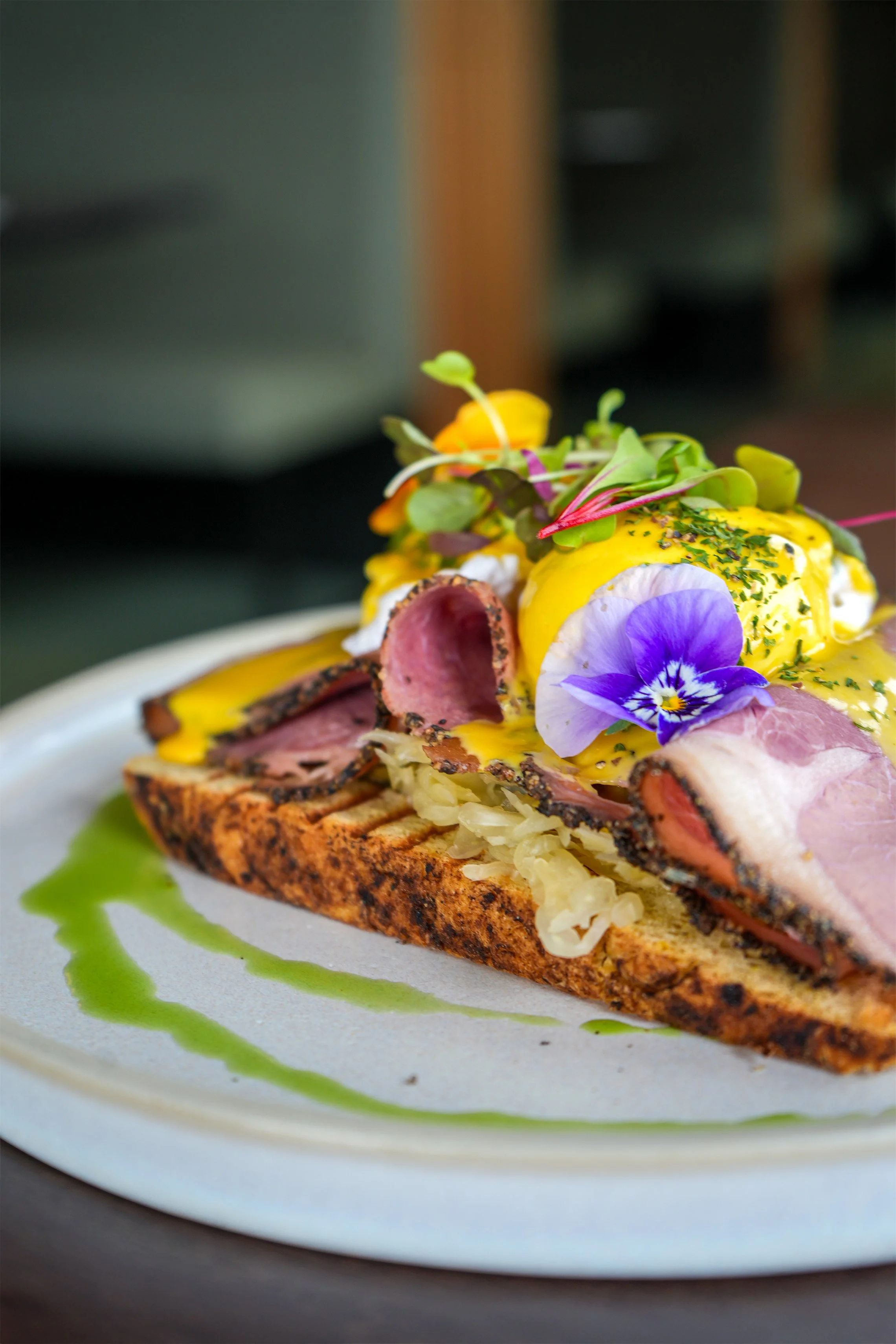 Open-faced sandwich with grilled bread, sliced roast beef, lettuce, poached eggs topped with hollandaise sauce, edible flowers, microgreens, and green sauce on a white plate.
