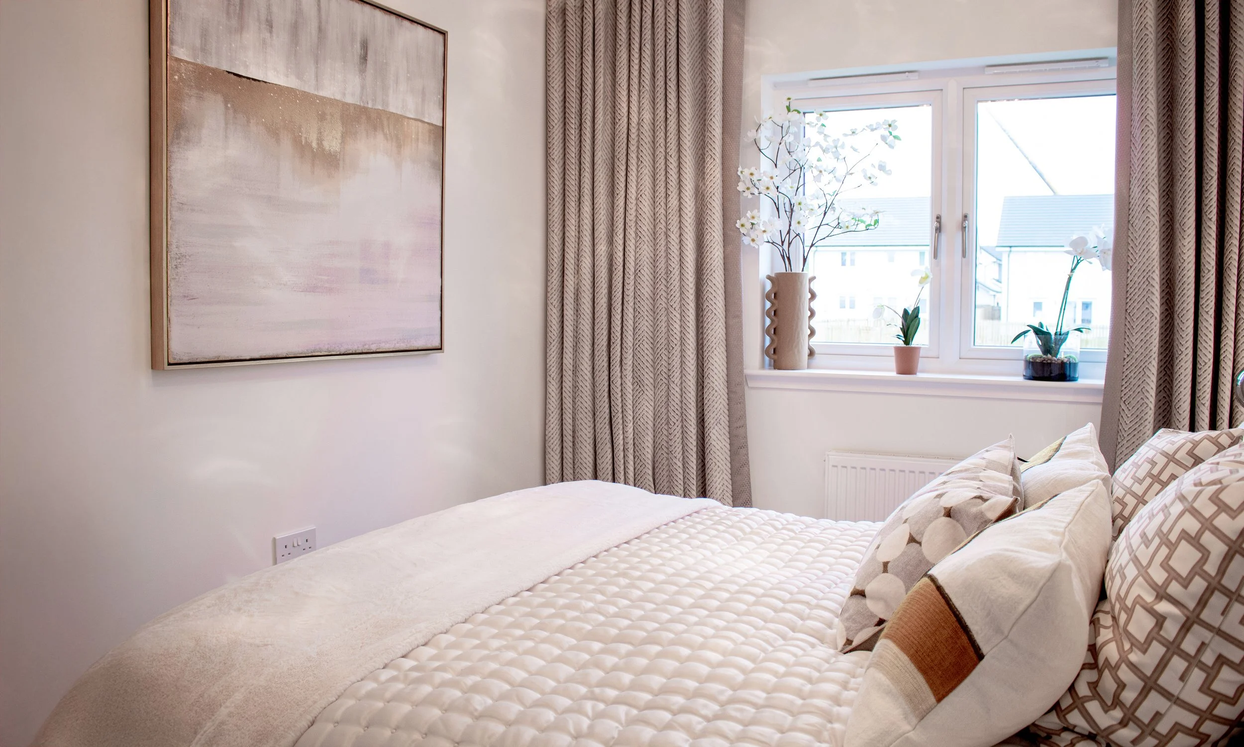 A cozy bedroom with a bed covered in a cream quilt and decorative pillows, a large abstract painting on the wall, and a window with beige curtains and potted orchids and plants on the windowsill.
