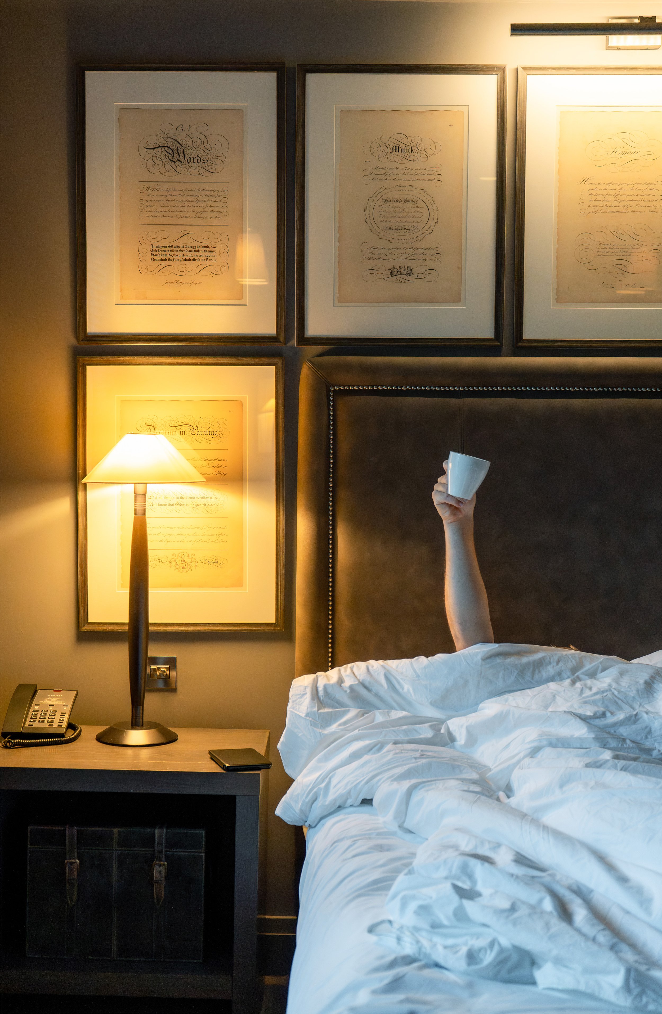 A person lying in bed holding a coffee cup, with a lamp illuminating the bedside table and framed documents on the wall behind.