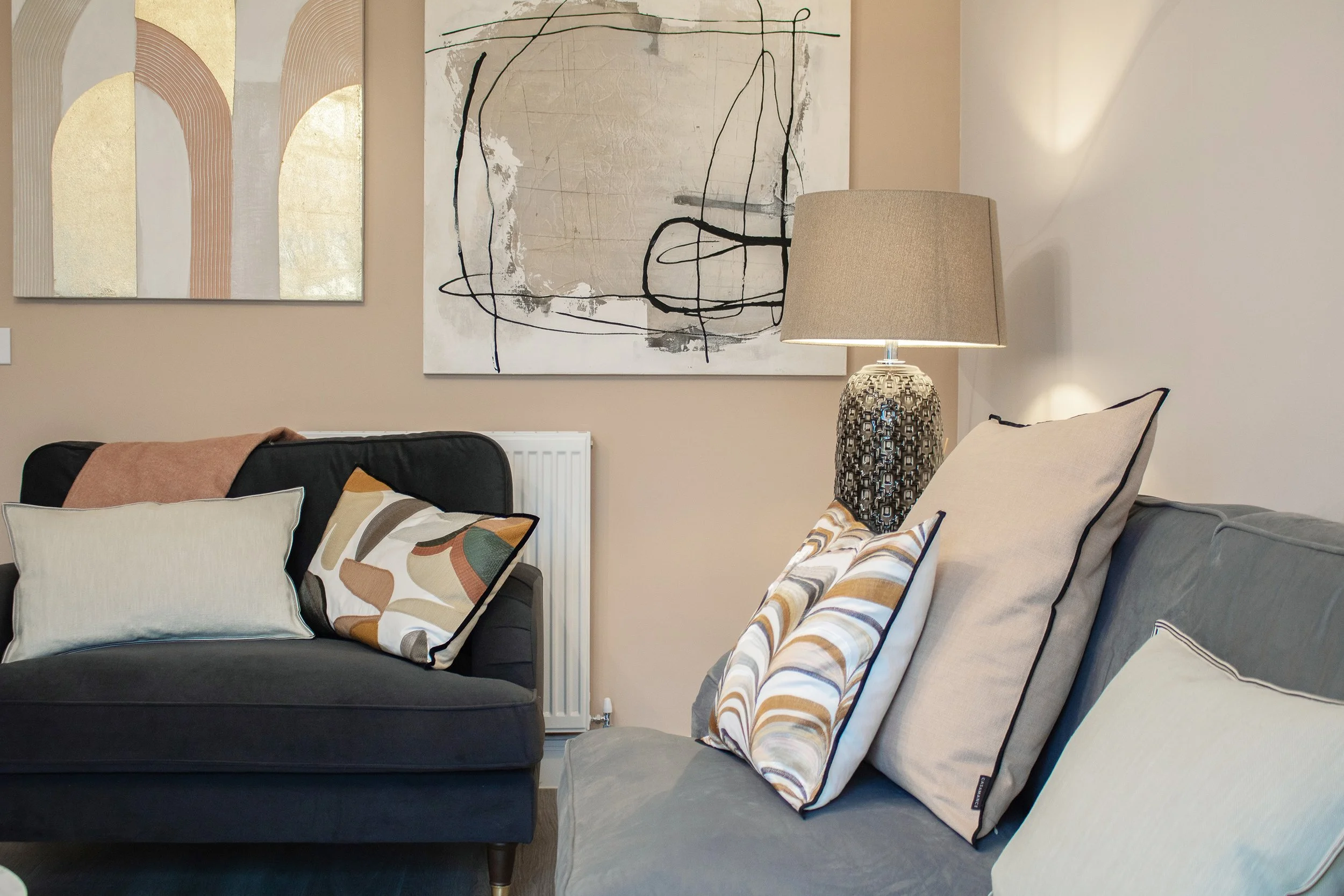 Living room with gray and black sofas decorated with cream and patterned pillows; wall art featuring abstract paintings; a table lamp on the side table; neutral-colored walls and a radiator below the artwork.