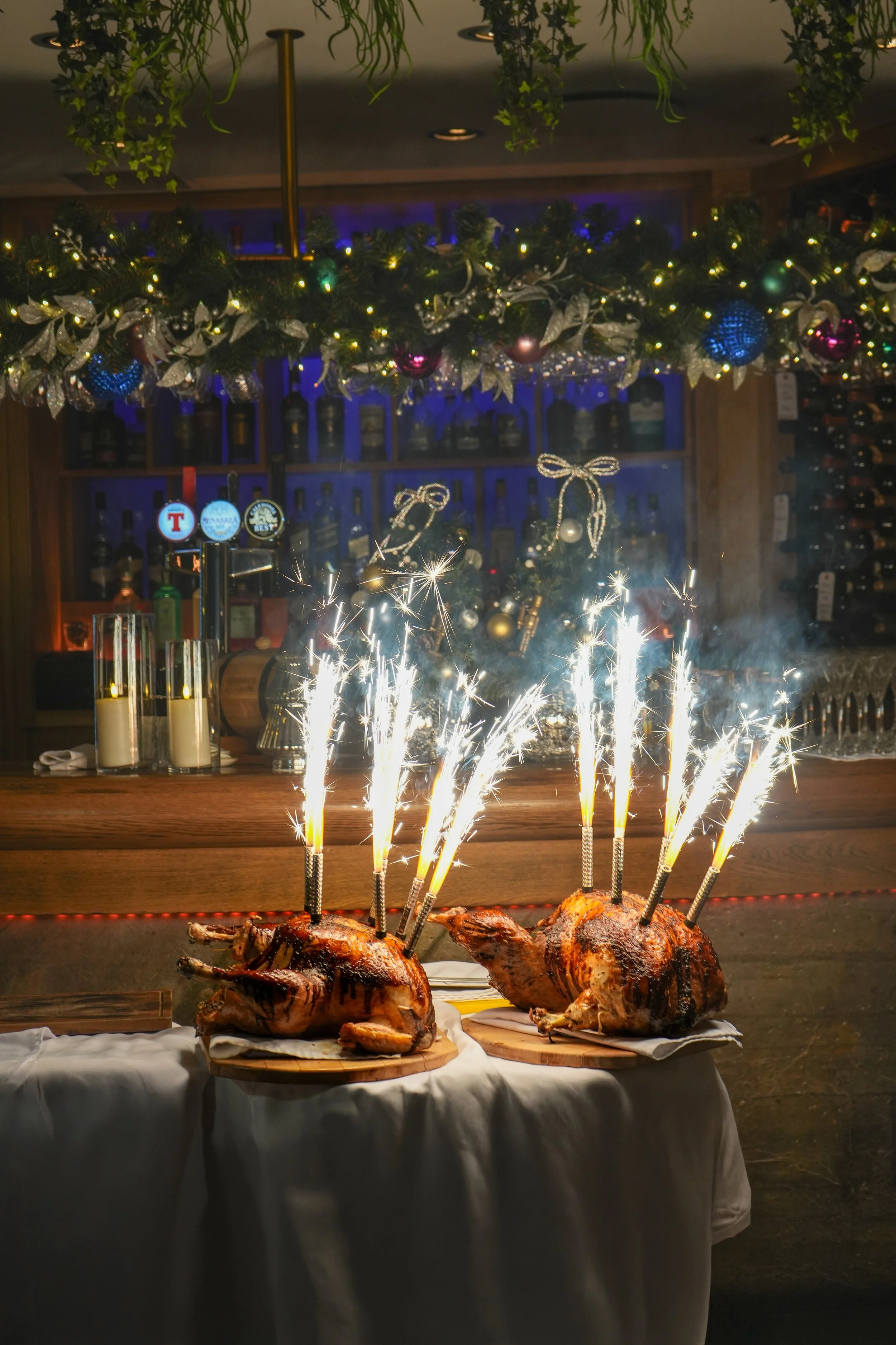 Two roasted chickens with sparklers on top, placed on wooden boards on a table decorated for a celebration, with Christmas decorations and bottles of alcohol in the background.
