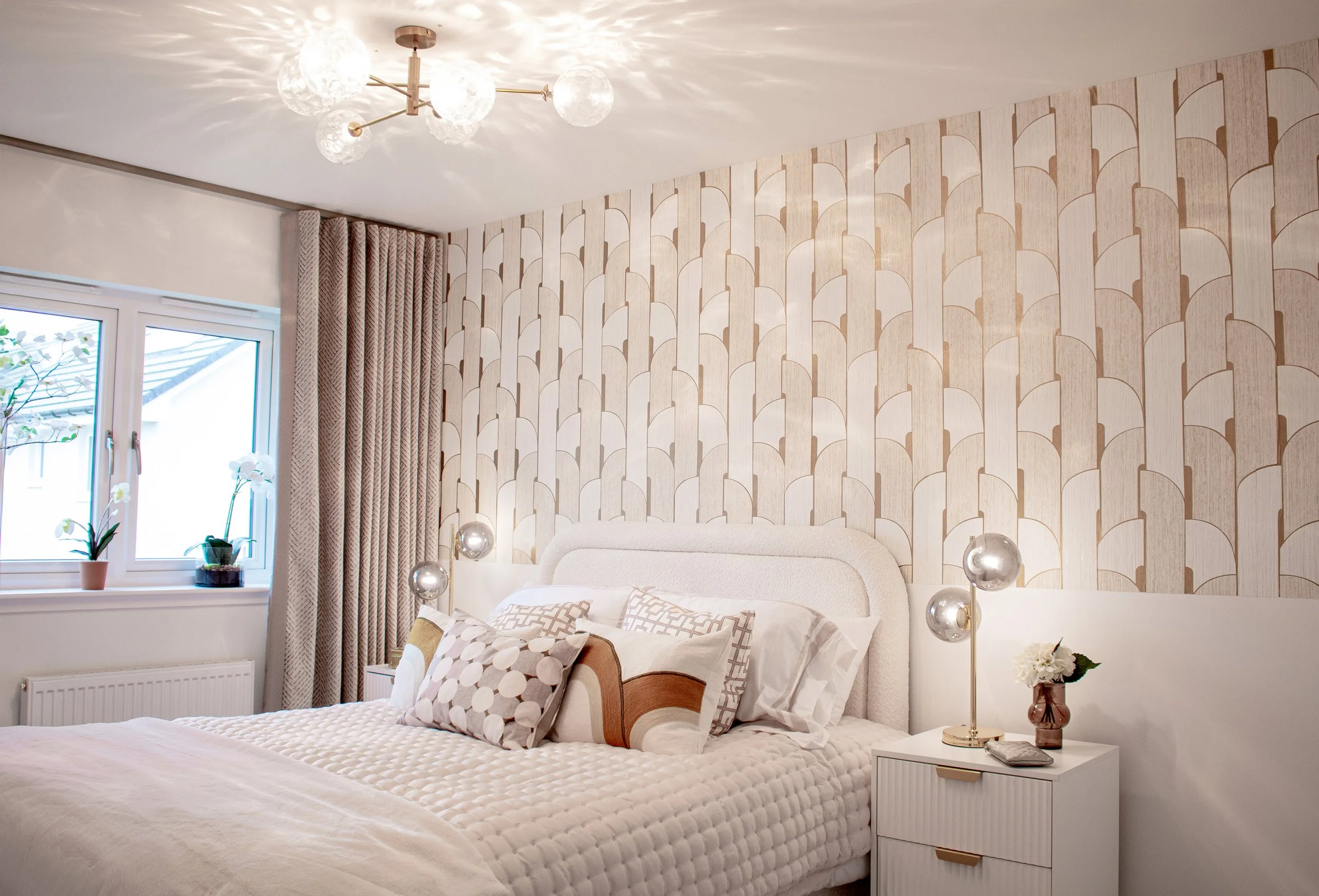 A bedroom with a bed against a patterned accent wall, white bedside tables with gold accents, a modern chandelier, and a window with beige curtains and potted plants.