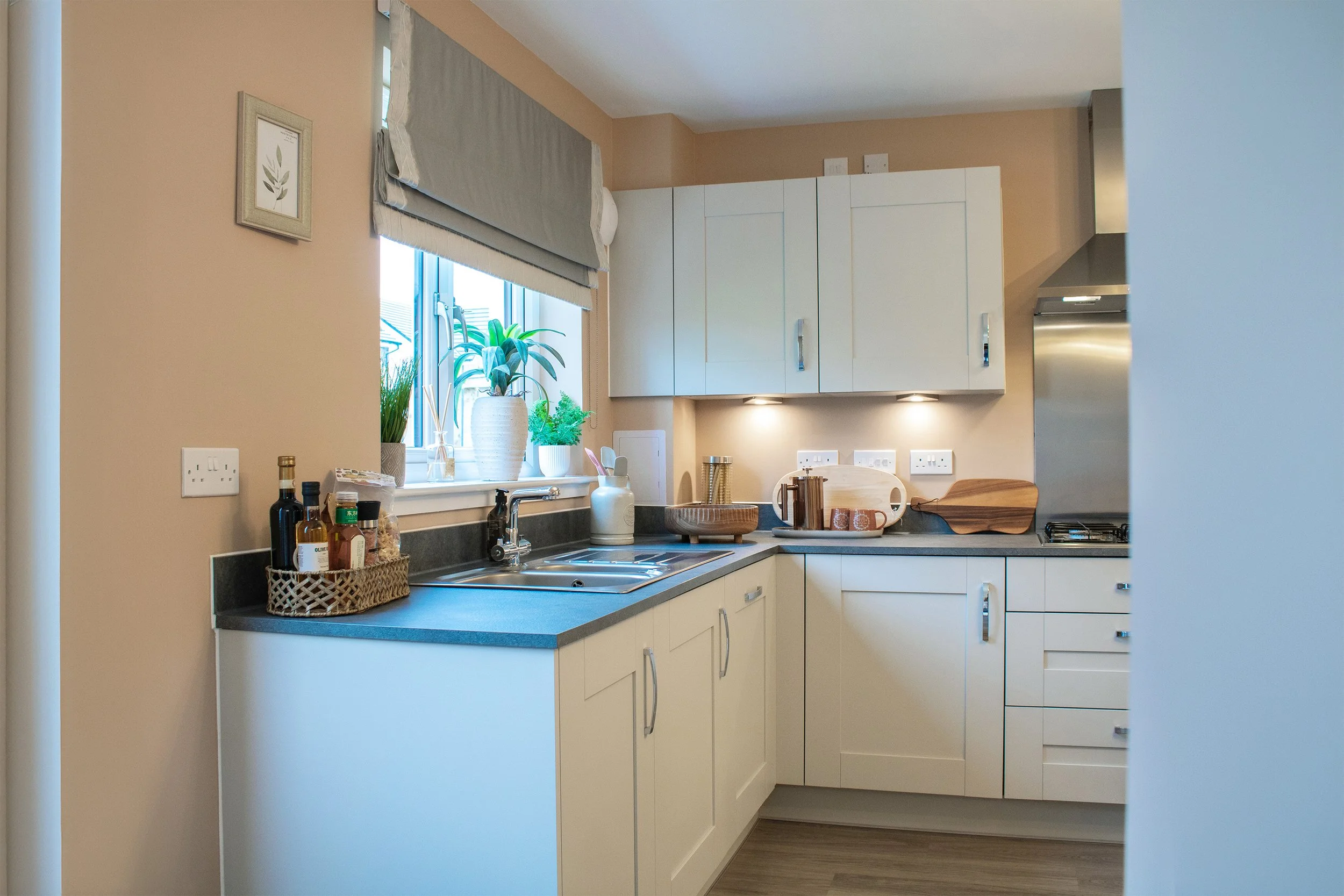 real estate photography of a modern Kitchen with cream cabinets, black countertop, and on the window ledge are potted plants. There are bottles and jars on the counter, a sink, and a window with a beige curtain.