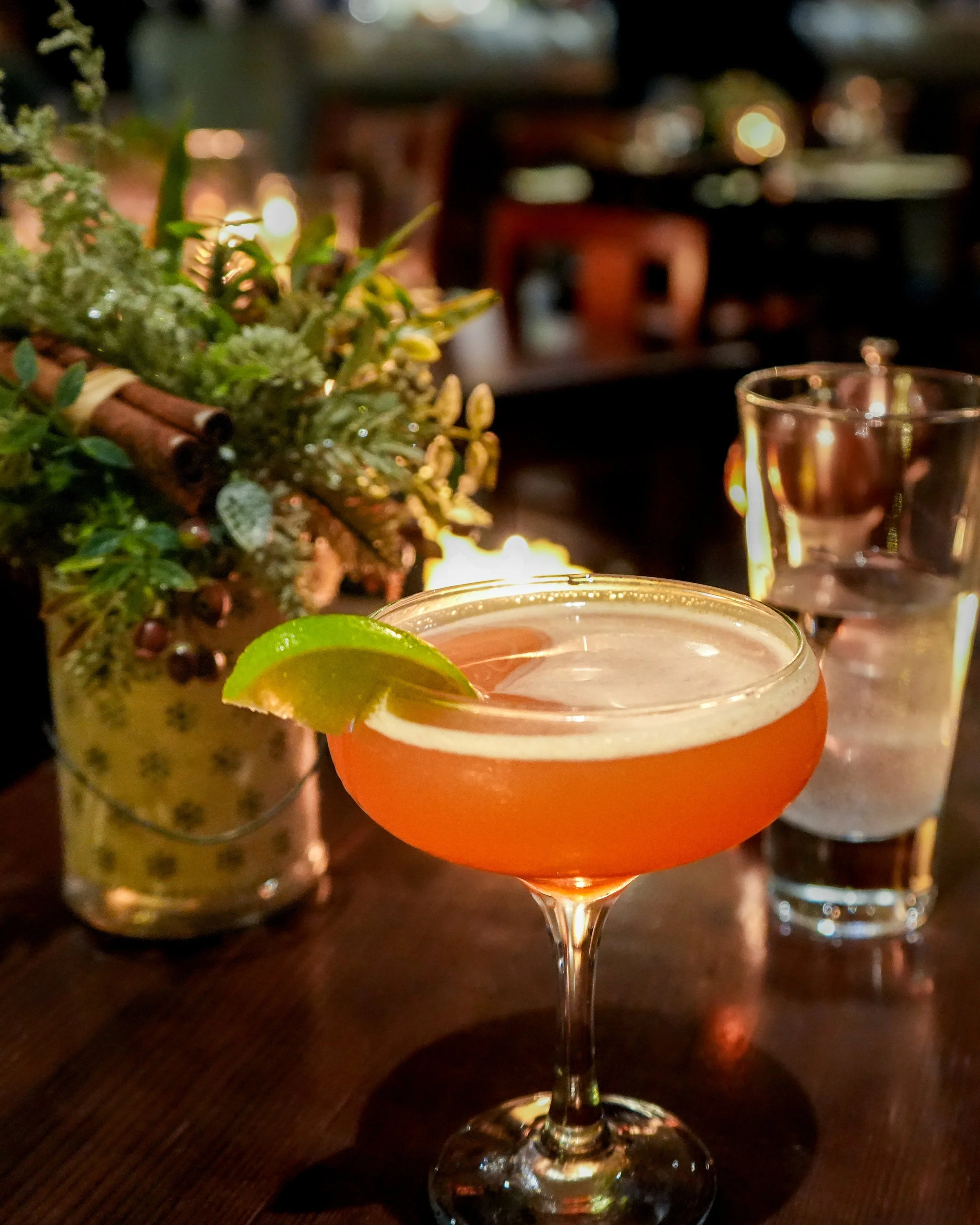 A cocktail in a coupe glass with a lime wedge garnish, set on a wooden table with a blurred background of a bar or restaurant, with a decorative greenery centerpiece and two glasses of water behind it.