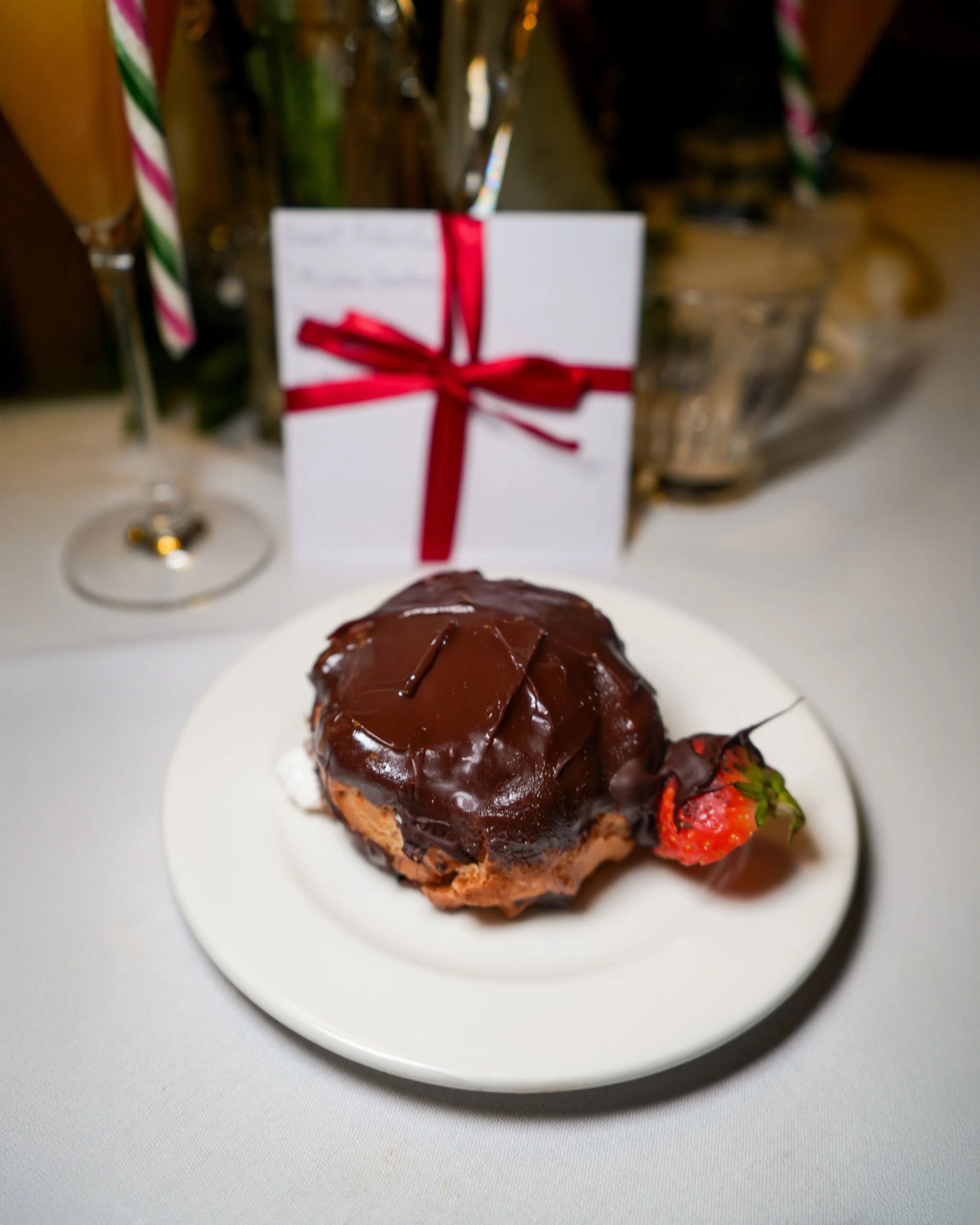 Chocolate-covered pastry with a strawberry on the side, on a white plate, with a white gift card tied with a red ribbon in the background.