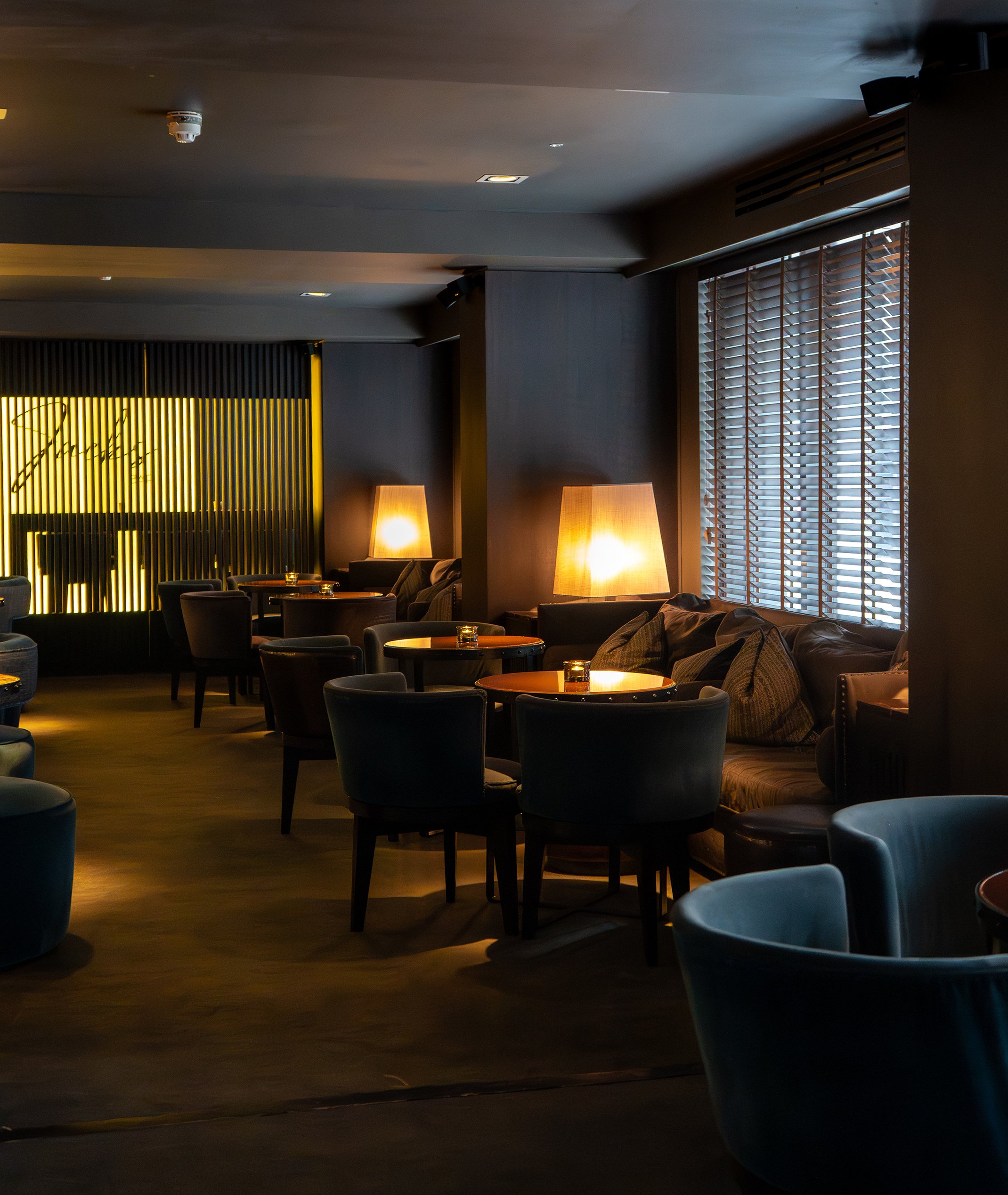 A dimly lit lounge with multiple tables and chairs, soft lighting from lamps, and window blinds.