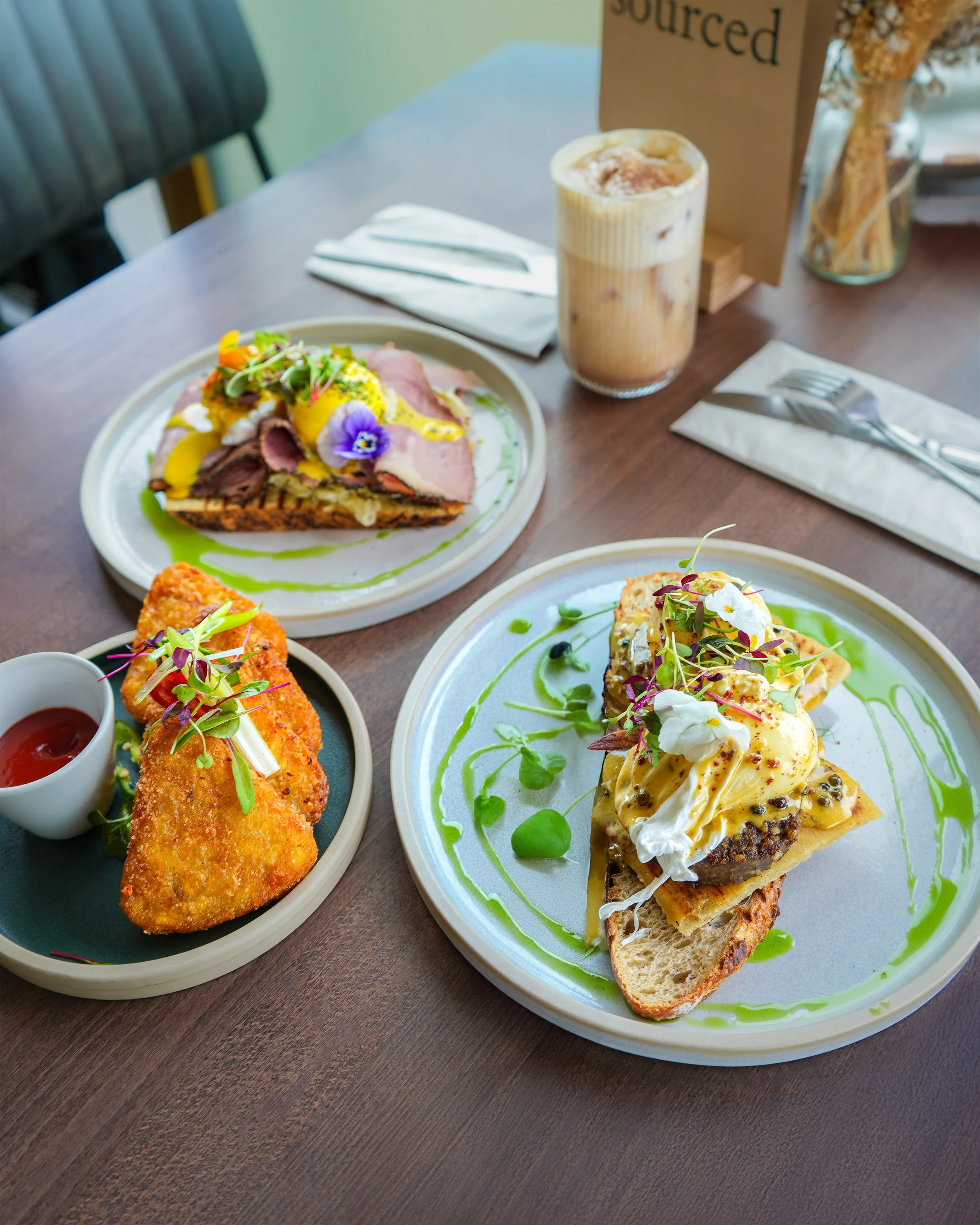 Three plates of food and a drink on a wooden table, with a fork and knife on napkins. The dishes include a savory tart with microgreens and flowers, a fried item with sauce and microgreens, and an open-faced sandwich with an egg, microgreens, and sauce.