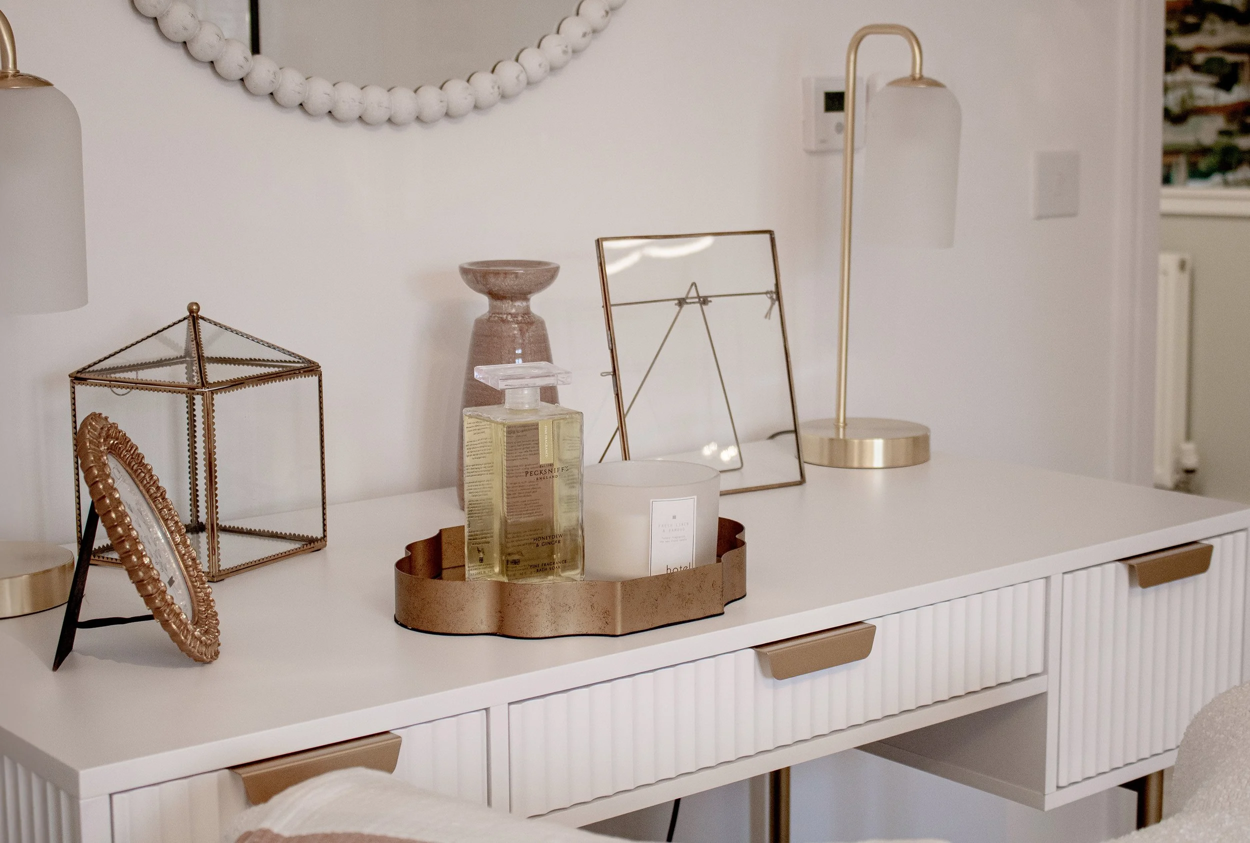 Decorative items on a white sideboard, including a gold table lamp, framed mirror, glass container with liquid soap, gold geometric terrarium, small photo frame, and a white candle, with a round mirror on the wall above.