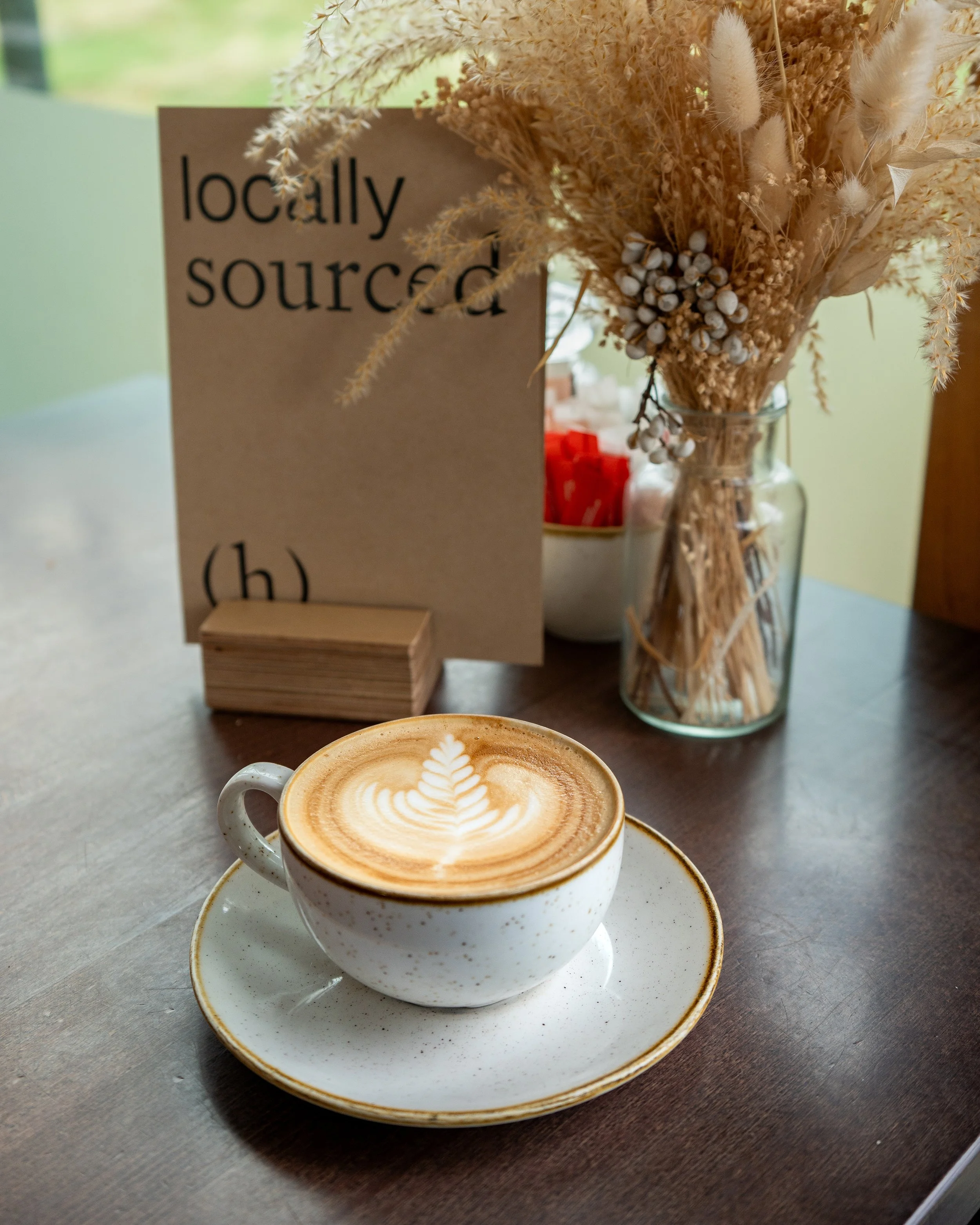 A cup of latte with leaf latte art on a wooden table, with a dried flower arrangement, and a sign that says 'locally sourced' in the background.