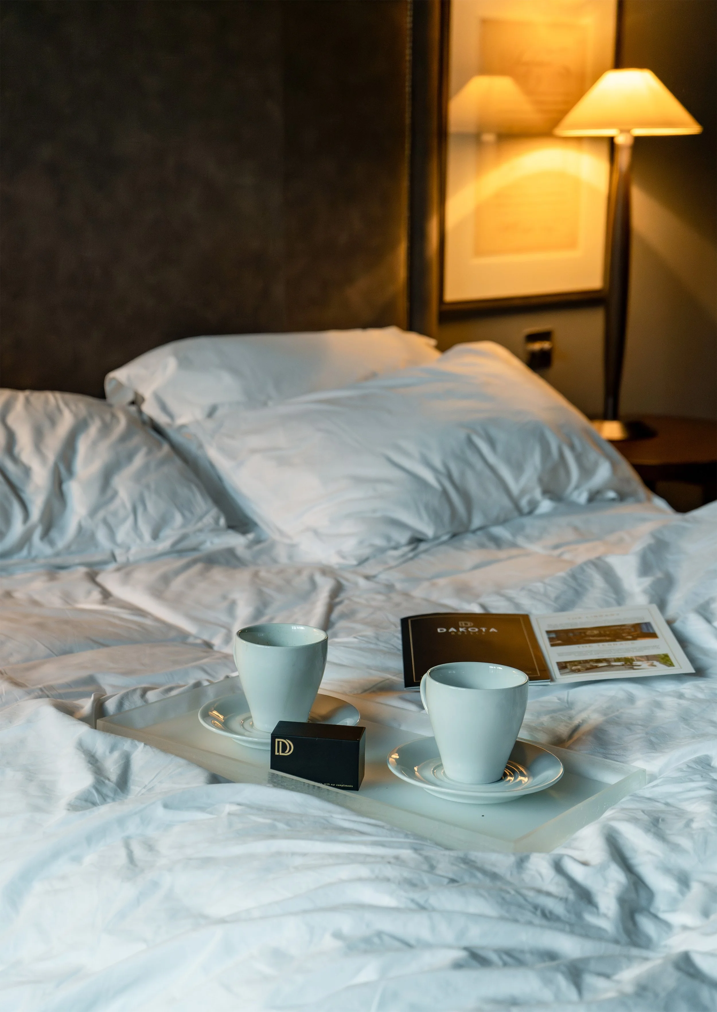 A luxury hotel bed with white sheets and pillows, a tray holding two white coffee cups on saucers, and an open brochure laying on the bed, with a bedside lamp illuminating the scene.