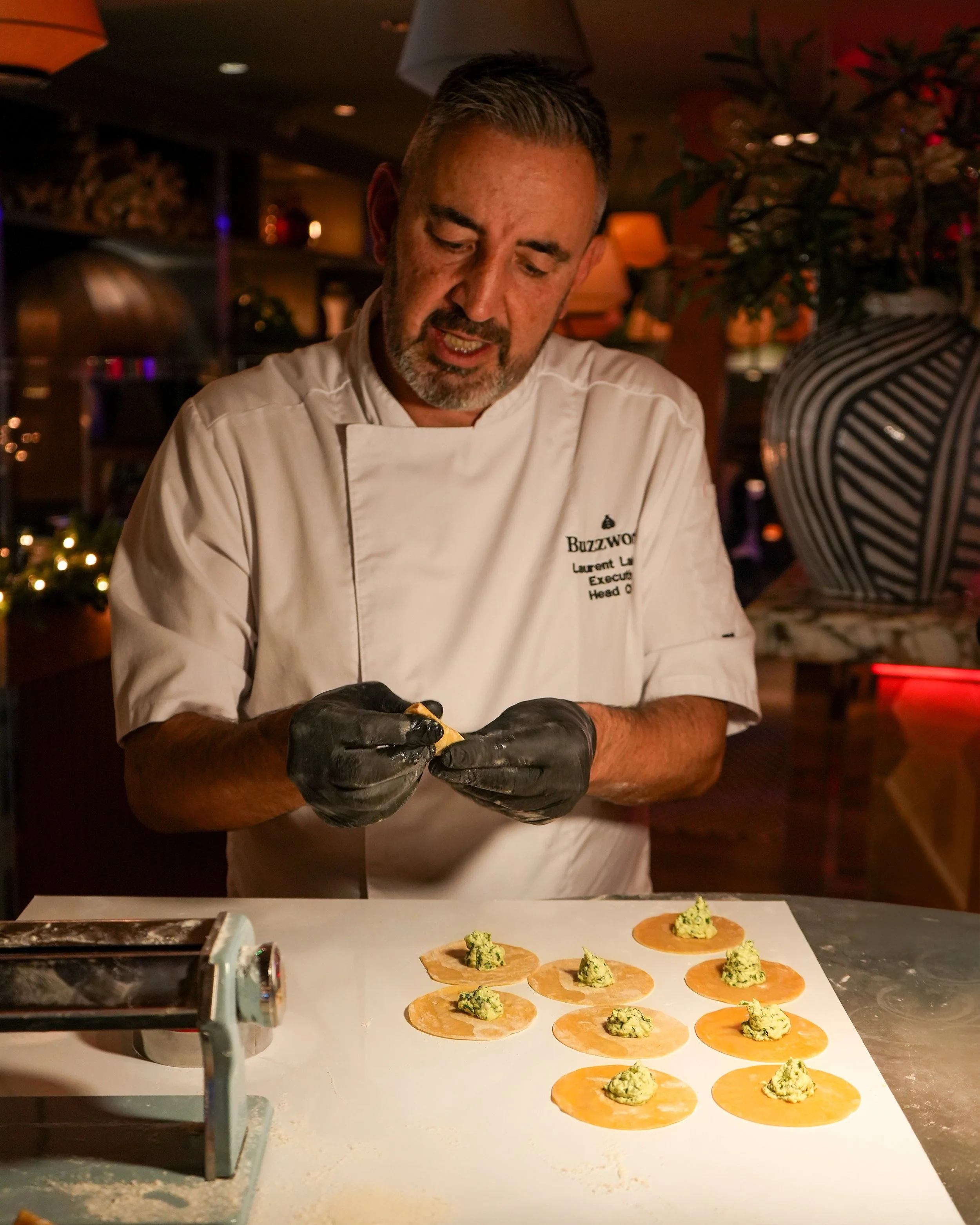 A chef wearing a white uniform and black gloves preparing small servings of food on a white surface, with a large decorative vase and plants in the background.