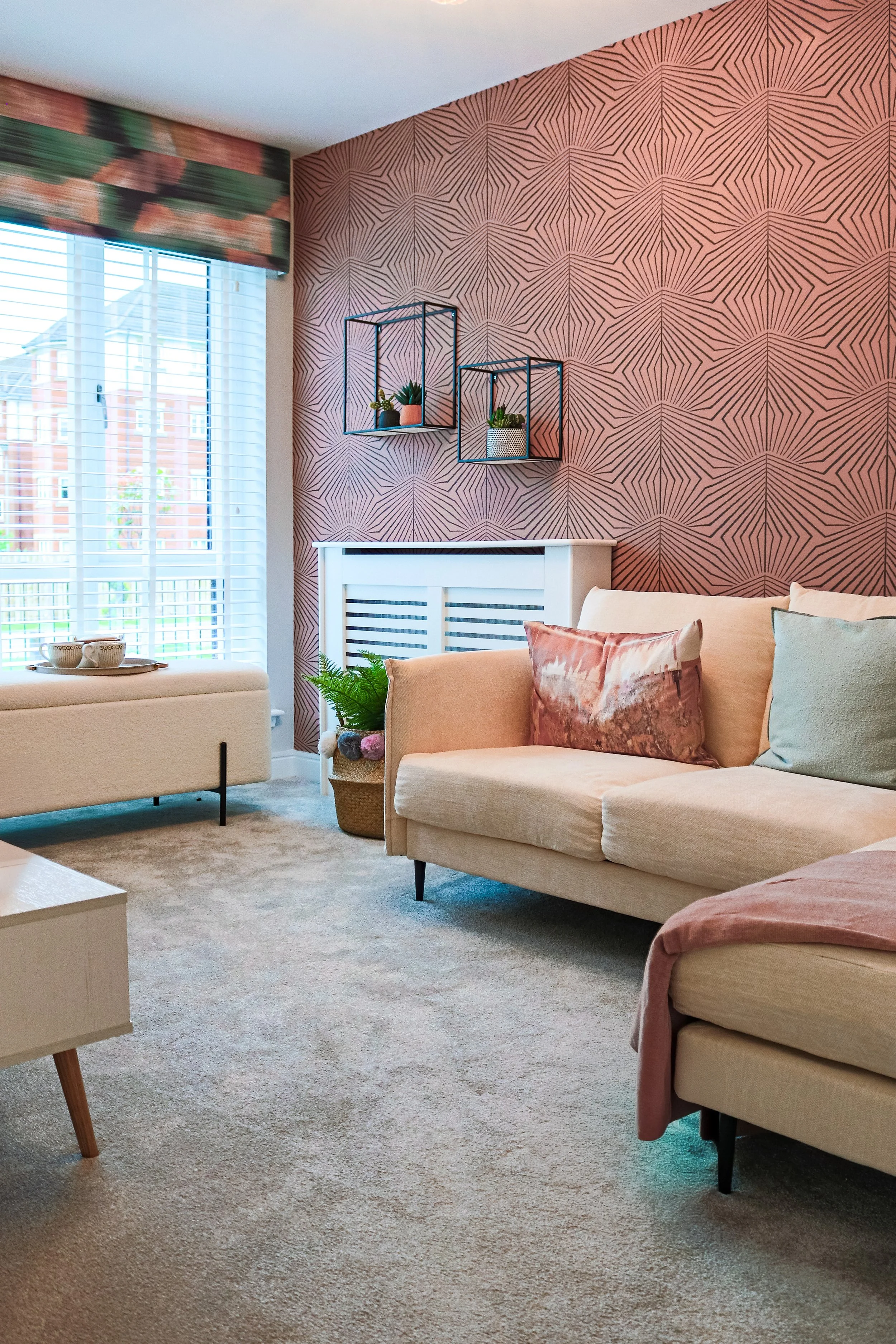 Living room with beige sofas, decorative cushions, a wall-mounted metal shelf with plants, a window with blinds and floral valance, patterned pink wallpaper, a white radiator cover, and a woven basket with ferns and pom-pom trim.