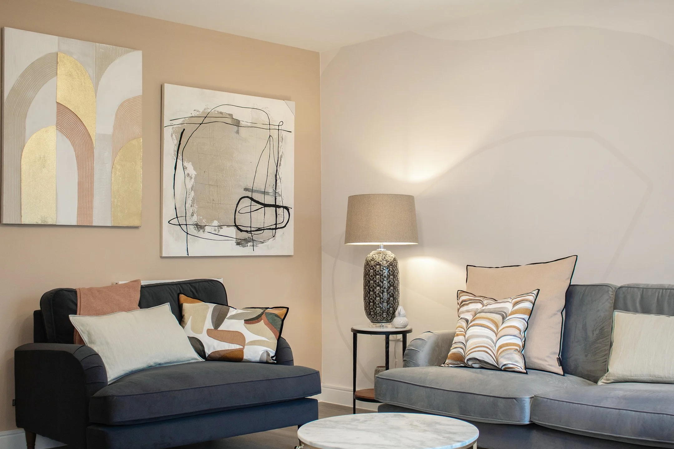 Living room with two sofas, pillows, artwork on beige walls, a lamp on a side table, and a round coffee table.