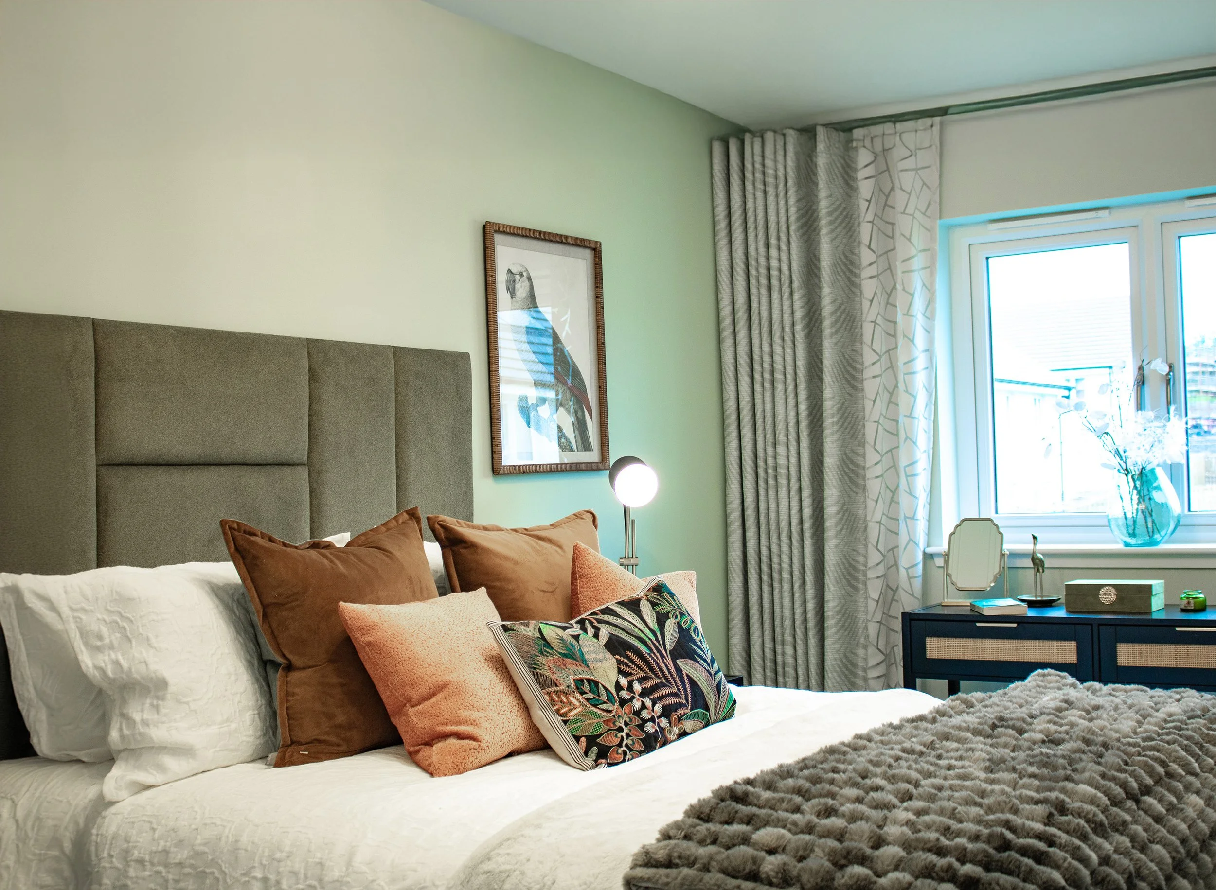 A neatly made bed with multiple pillows in front of a green wall, with a framed parrot picture, beside a window with curtains, a bedside lamp, and a navy dresser with decor in a bright bedroom.