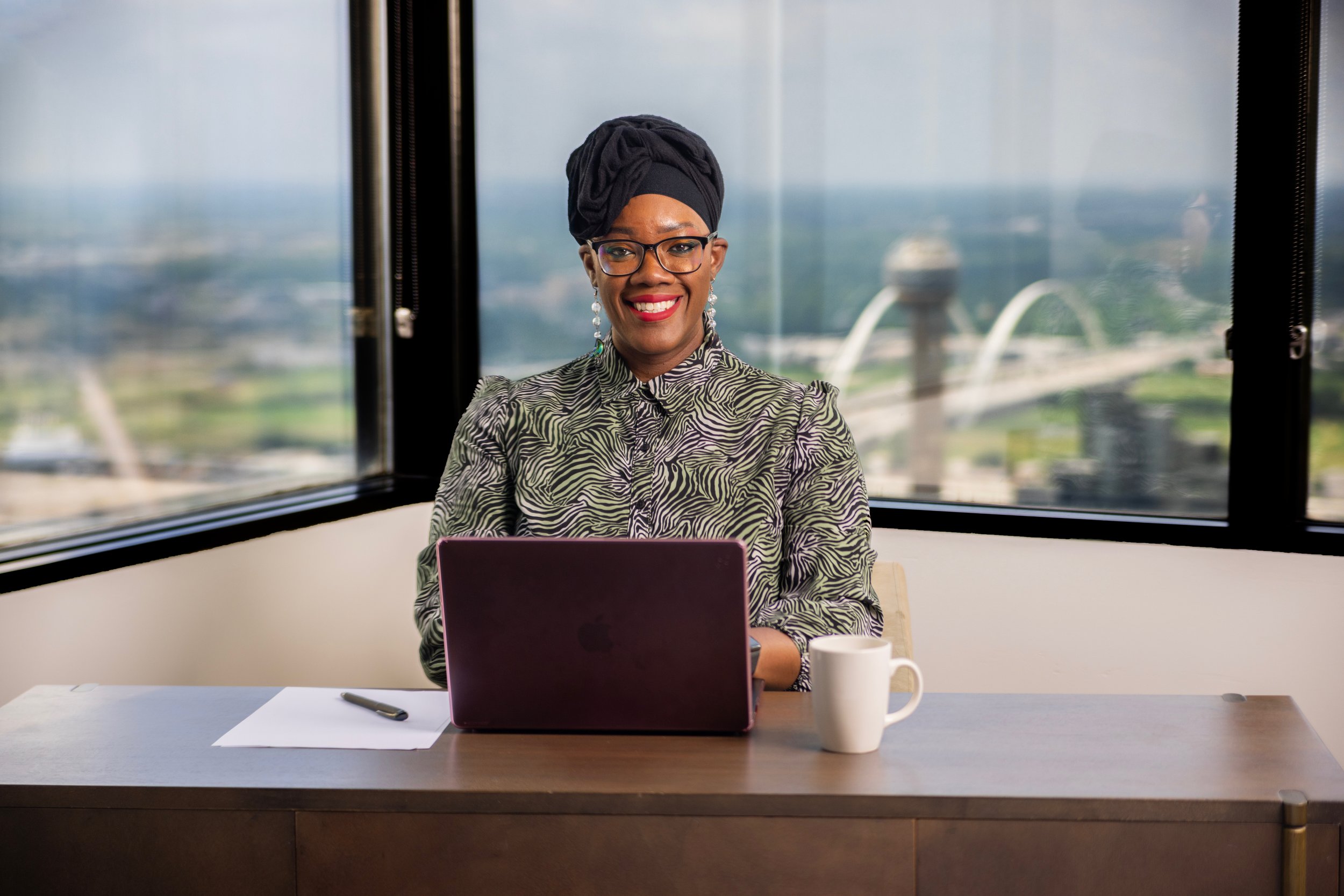 Smiling woman sitting at a desk with a laptop, papers, pen, and white mug in front of a large window with a cityscape view, including the Space Needle.