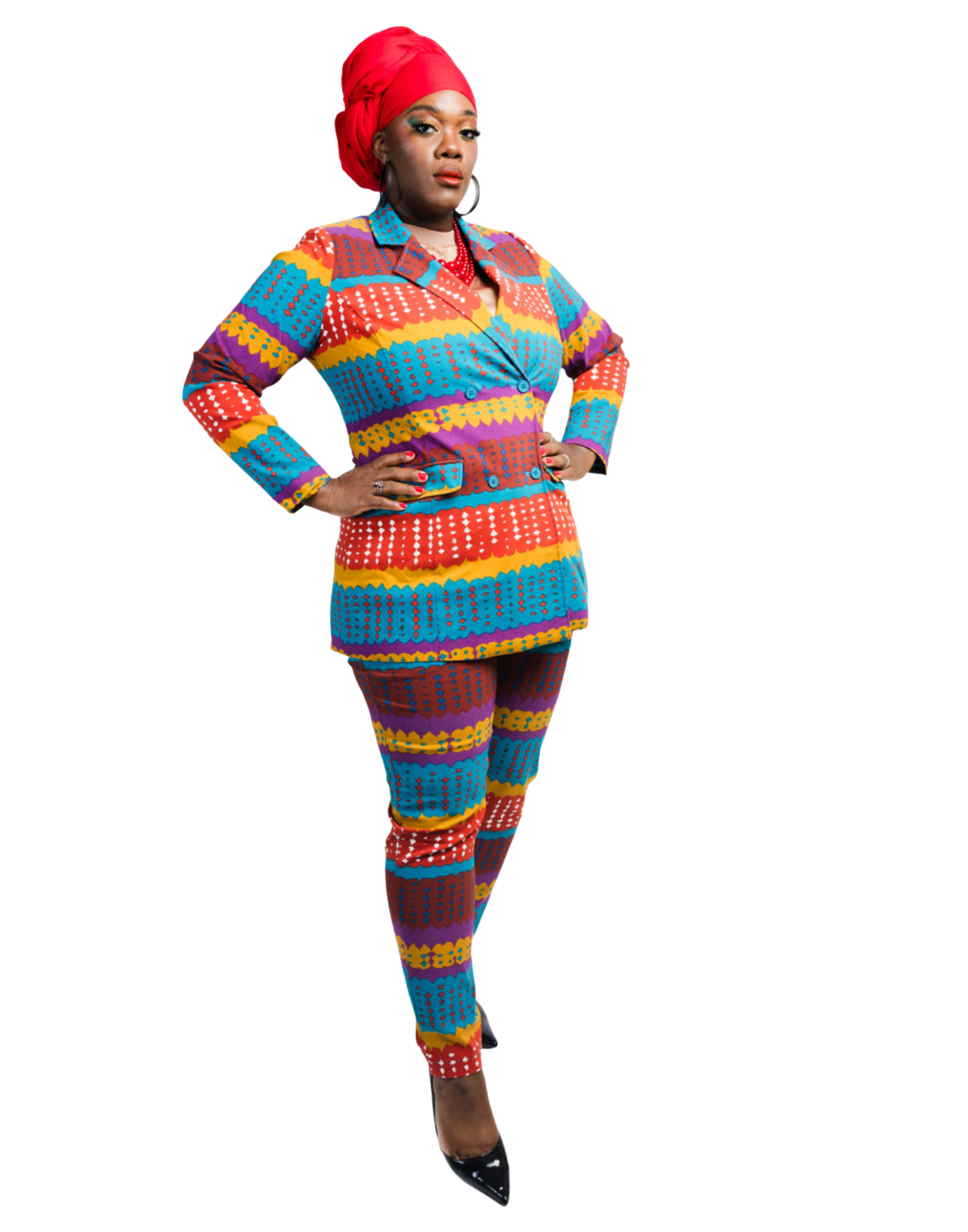 A woman dressed in a colorful, patterned suit with red, blue, yellow, purple, and white designs, wearing a red headwrap, earrings, and a red beaded necklace, standing with hands on hips against a white background.