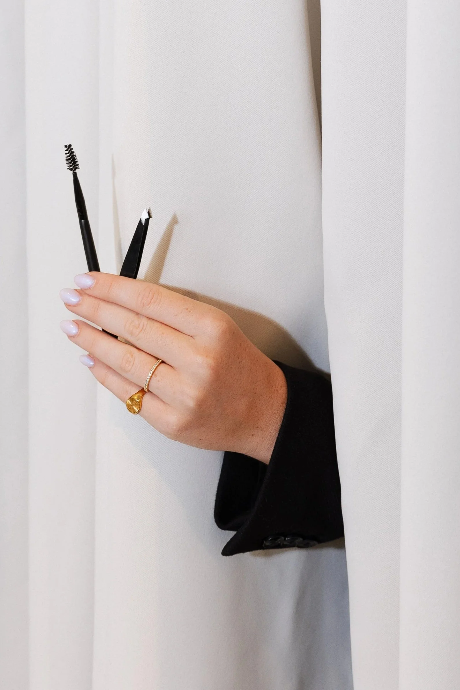 A hand with a gold ring peeking out from a white door window, holding two grooming tools: an eyebrow brush and tweezers.