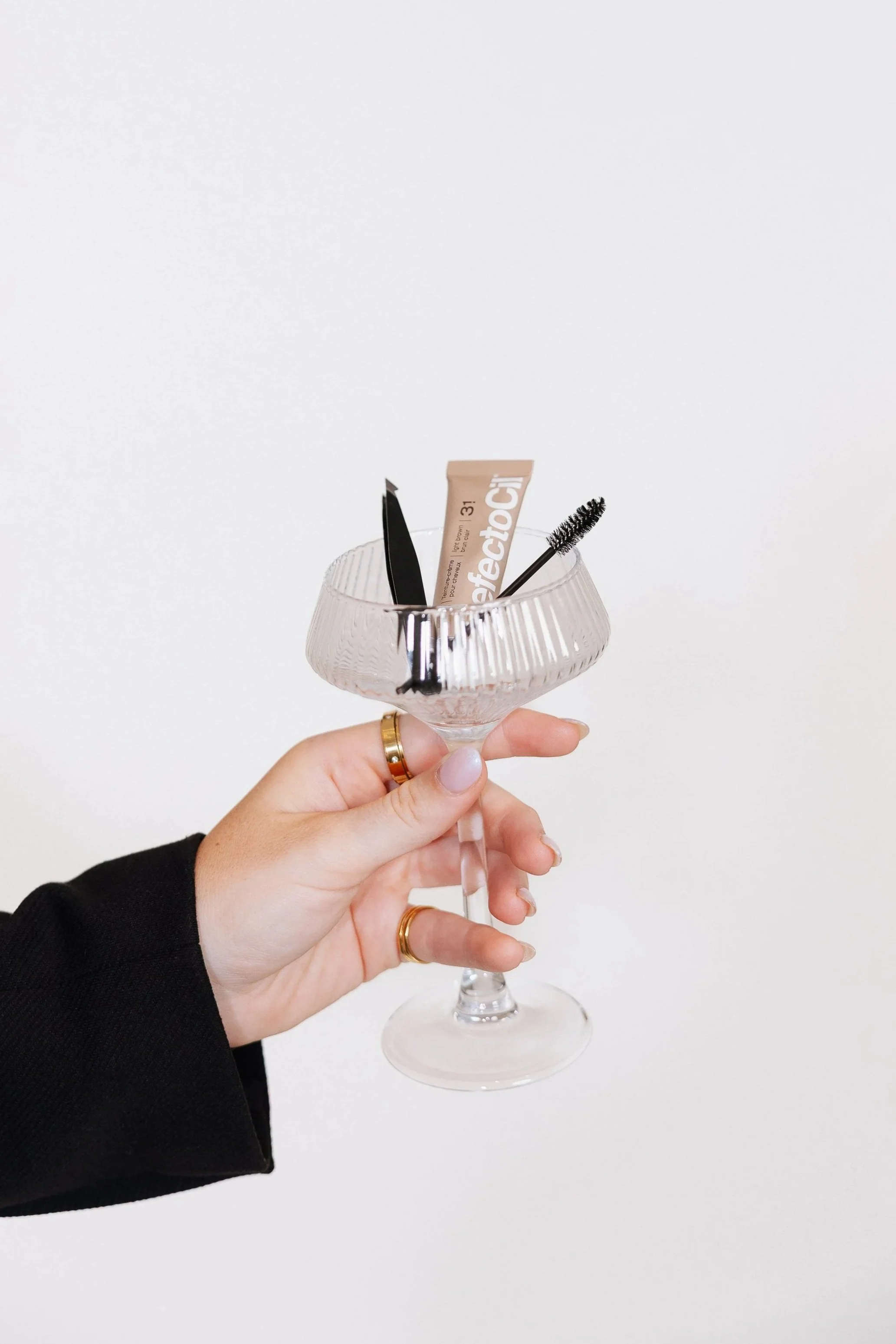 Hand holding a clear vintage-style glass cocktail coupe with a black manicure, gold rings, and a black sleeve, containing a tube of Fenty Beauty concealer, a black makeup brush, and a black mascara wand against a plain white background.