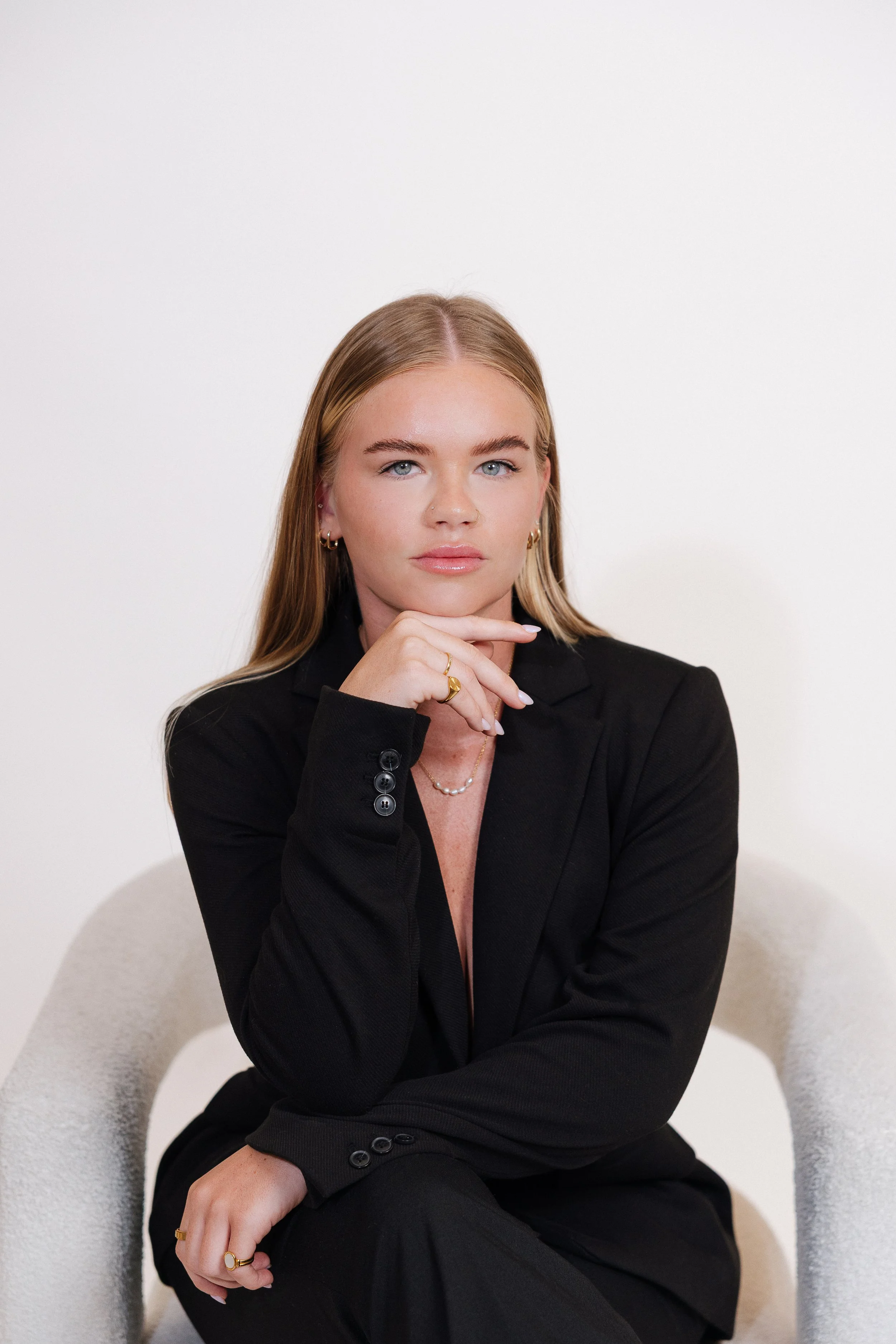 A young woman with blonde hair and blue eyes, dressed in a black blazer, sitting on a white chair against a plain white background.