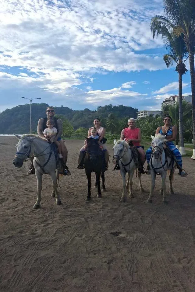 Four adults and two children riding horses on a dirt path in a tropical setting with palm trees, hills, and blue sky with clouds in the background.