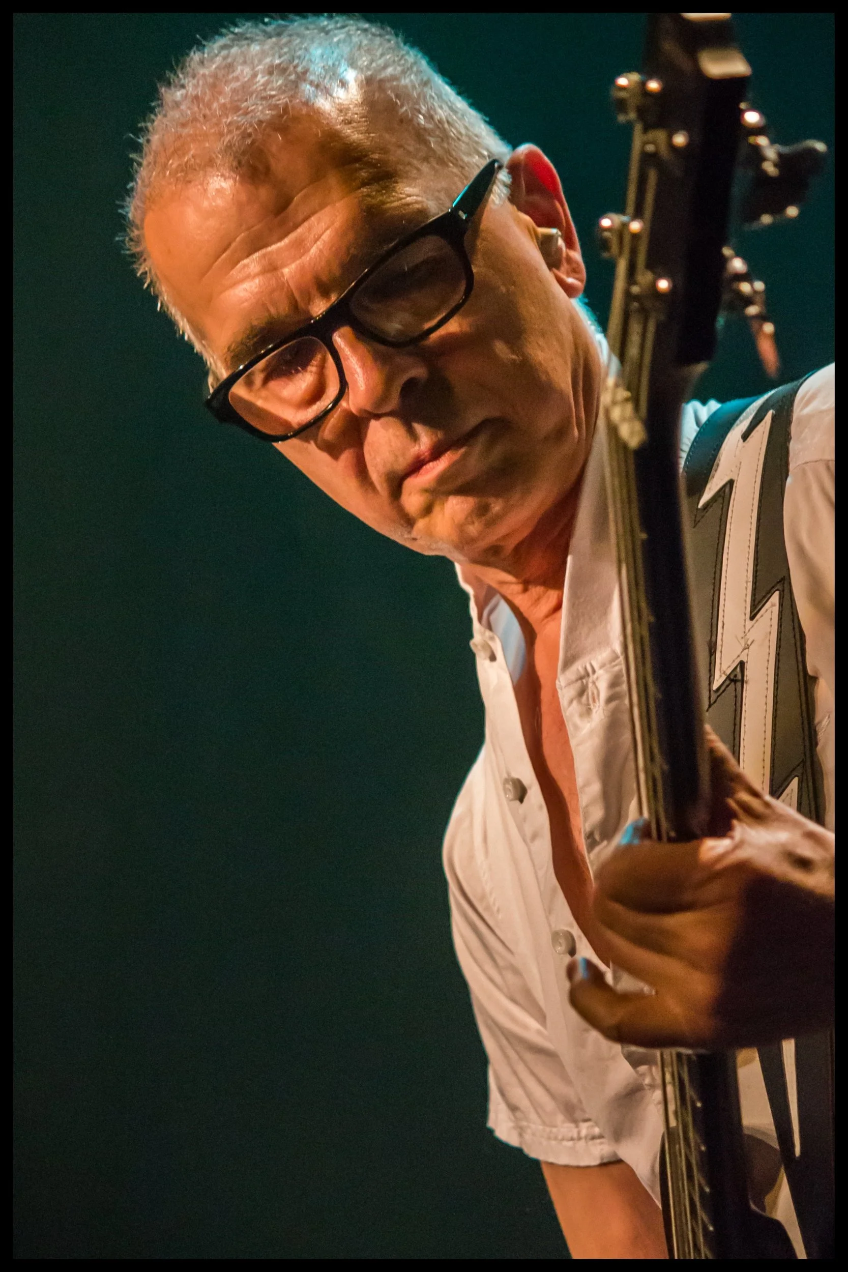 A close-up live concert portrait of legendary music producer and bassist Tony Visconti performing on stage, tilting his head with focused expression over his bass guitar under stage lighting. Expert live music photography capturing iconic classic roc