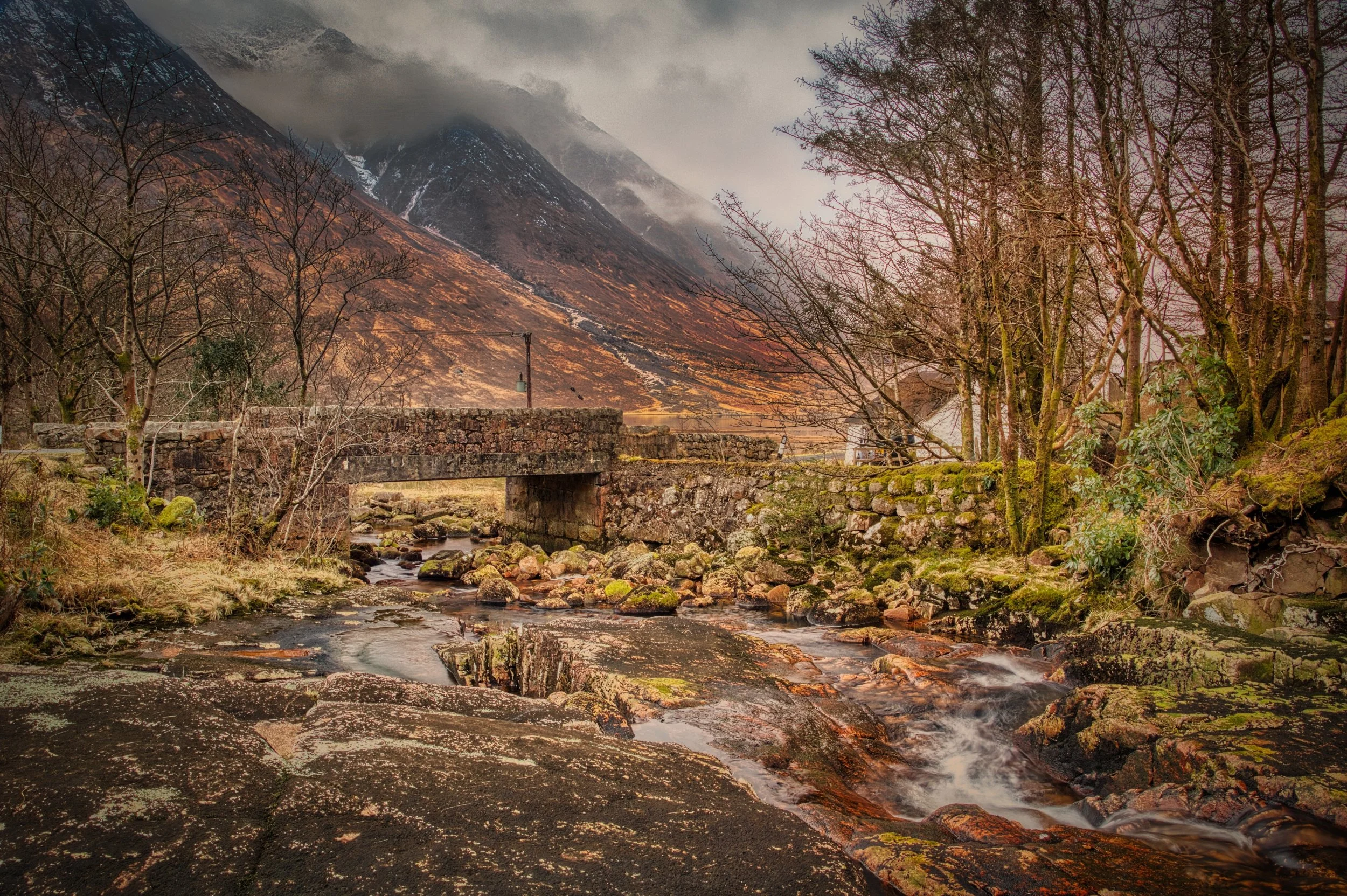 An old stone bridge crossing a rushing, rocky stream in Glen Etive, with rugged copper-toned mountain slopes disappearing into low, atmospheric mountain mist. Fine art Scottish landscape photography capturing the timeless textures and moody weather o