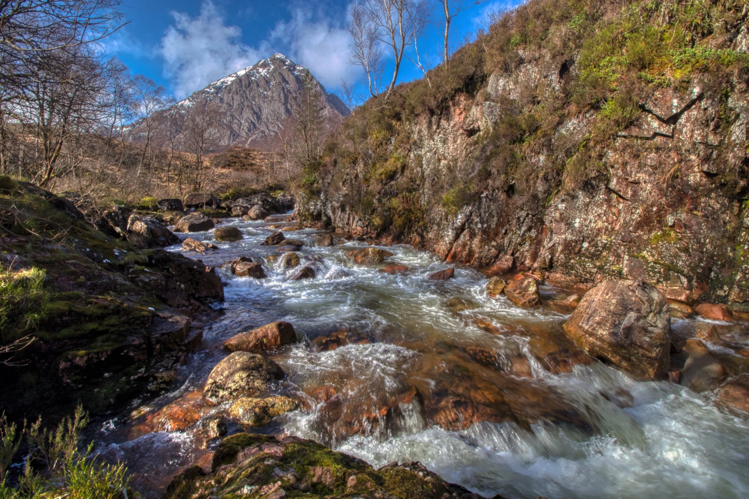 The rushing waters of the River Etive cascading over rocks, with the dramatic peak of Buachaille Etive Mòr rising sharply under a clear blue sky. Fine art Scottish landscape photography capturing the rugged stone textures and untamed beauty of Glenco