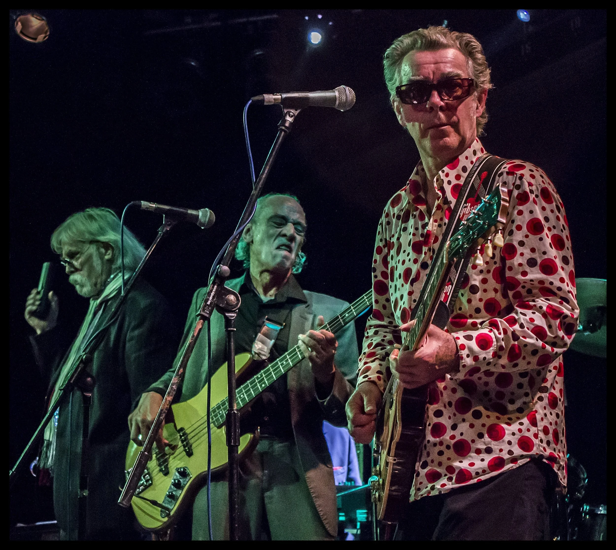 The Blockheads performing live on a dark stage, featuring guitarist Chaz Jankel in a red polka-dot shirt and bassist Norman Watt-Roy digging into a bass guitar under stage lighting. Energetic concert photography capturing the raw intensity of classic