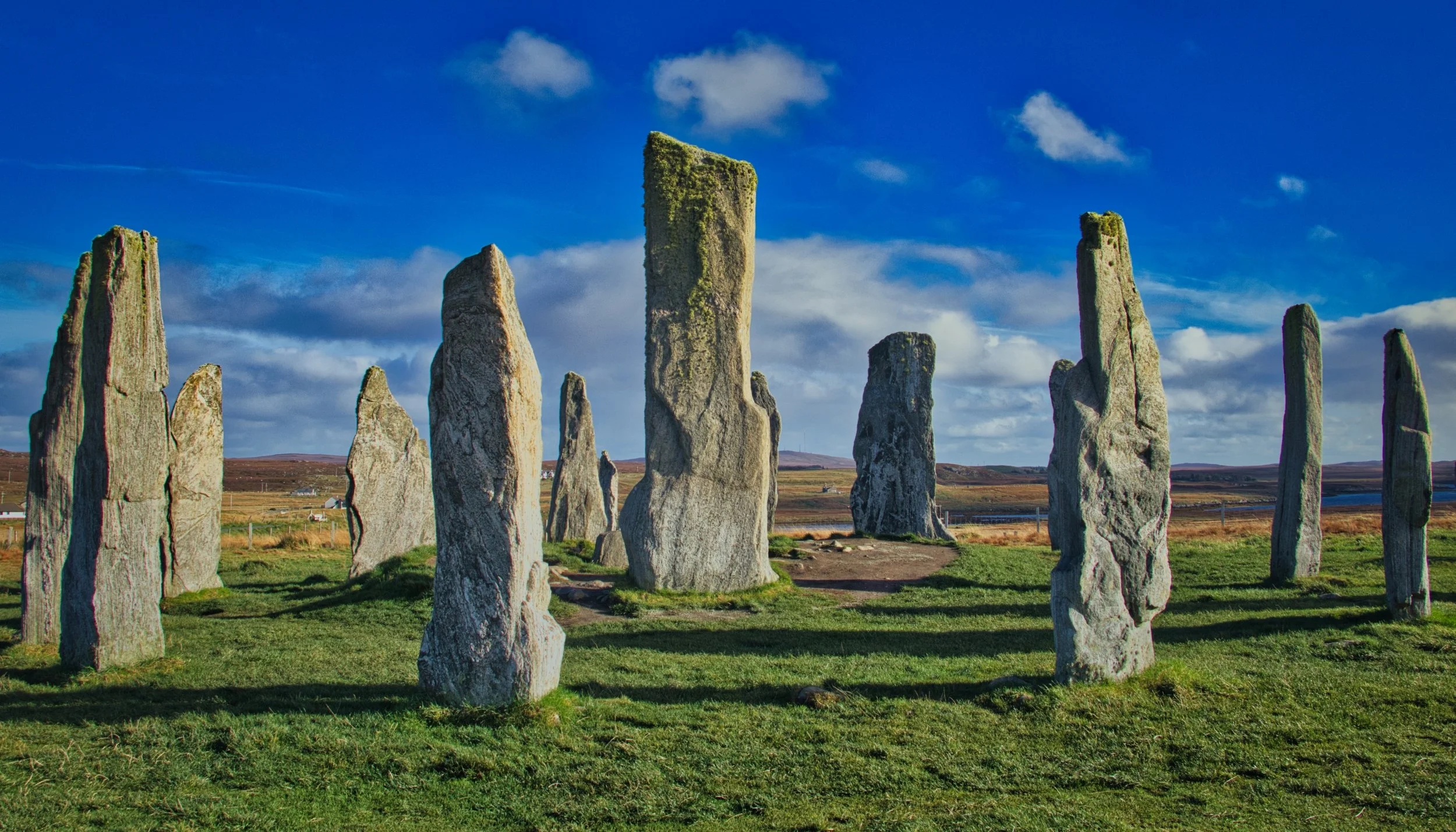 The ancient Lewisian gneiss of the Callanish Standing Stones on the Isle of Lewis, towering under a vibrant blue sky with dramatic scudding clouds. Landscape photography capturing the timeless scale and long, deep shadows of Scotland's historic monol