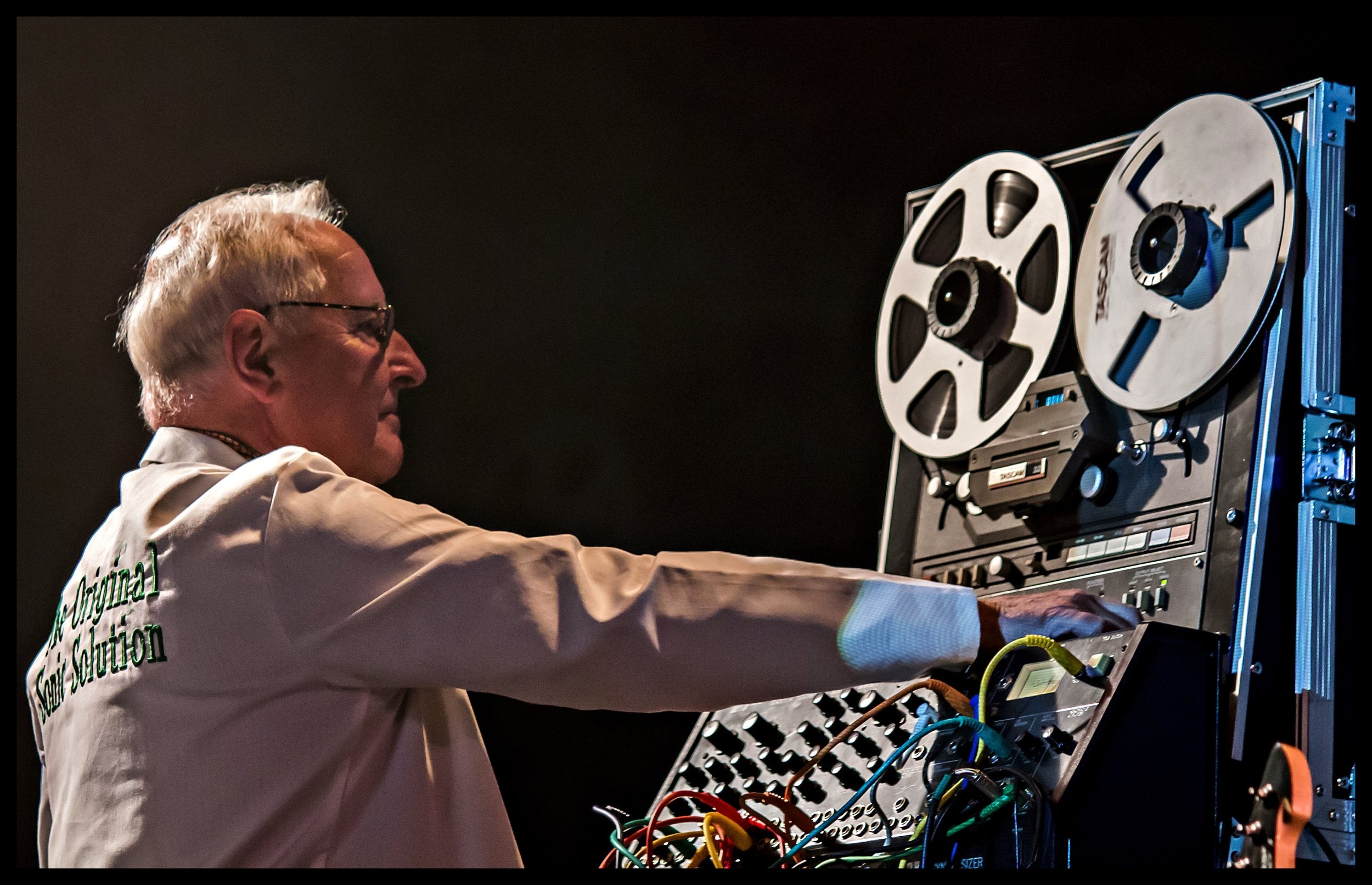 Legendary BBC Radiophonic Workshop composer Dick Mills operating a vintage Tascam reel-to-reel tape machine and synthesizer setup during a live performance. Specialized music photography capturing electronic audio pioneering and analogue studio gear.