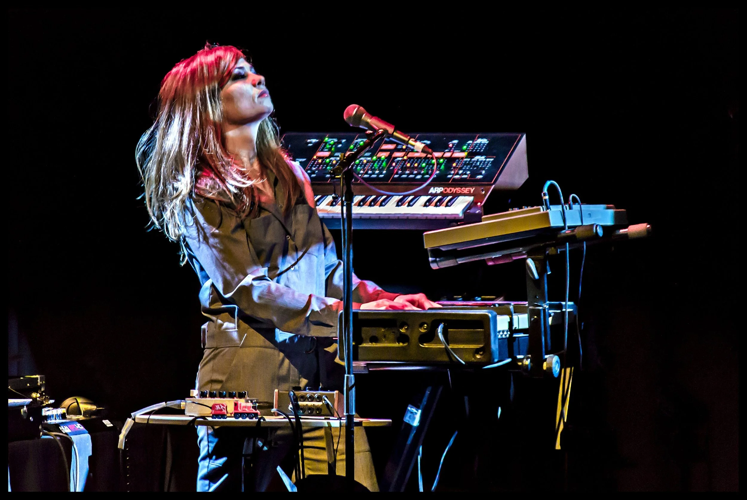 Musician and composer Tara Busch performing live, under the name of I Speak Machine, on a dark stage, operating a synthesizer setup with a vintage ARP Odyssey keyboard illuminated under stage lighting. Classic live music photography capturing artisti
