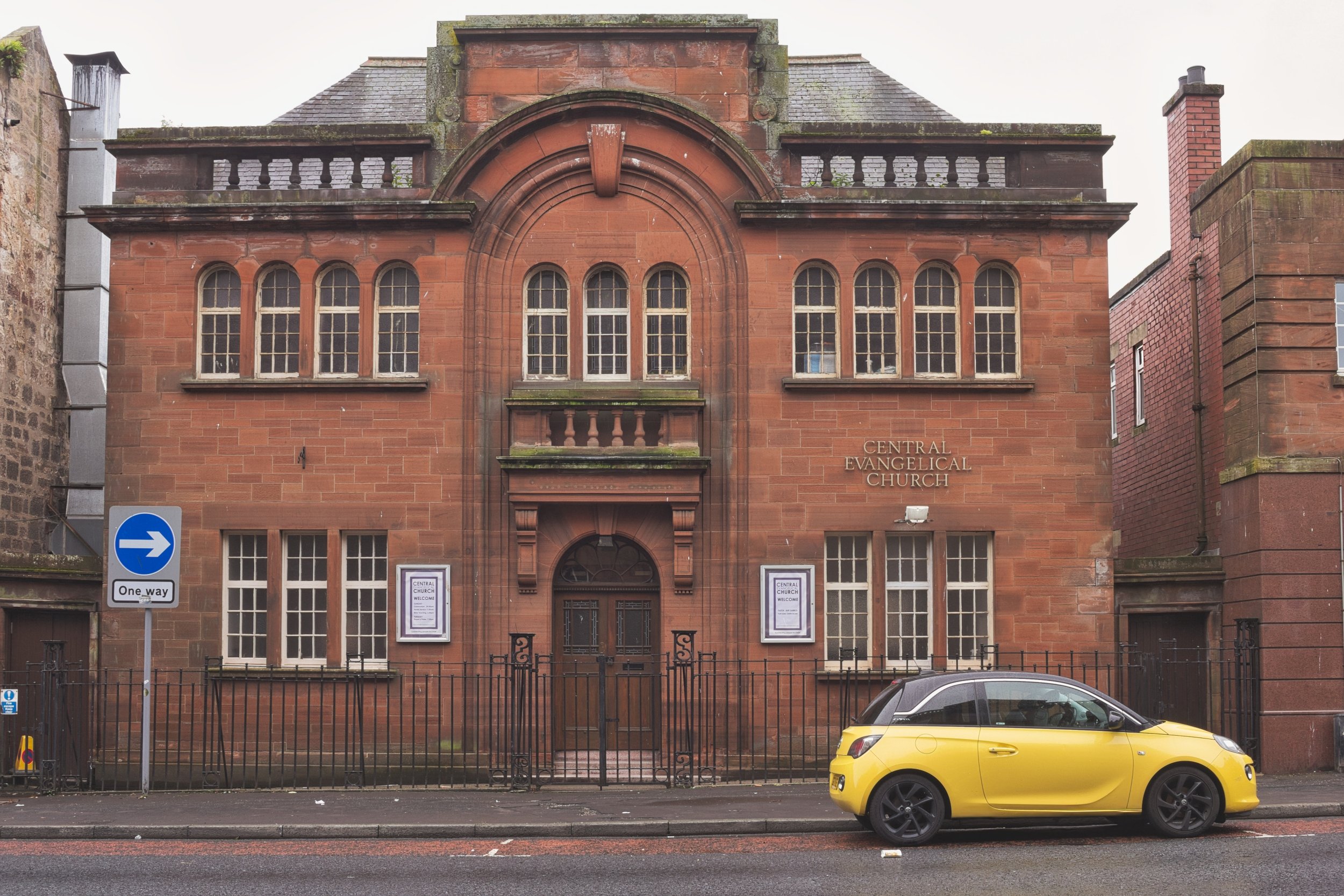 The red sandstone facade of the Central Evangelical Church fronted by black iron railings and a bright yellow car under an overcast sky in Kilmarnock, East Ayrshire. Deadpan urban landscape photography capturing the stark contrasts of everyday town a