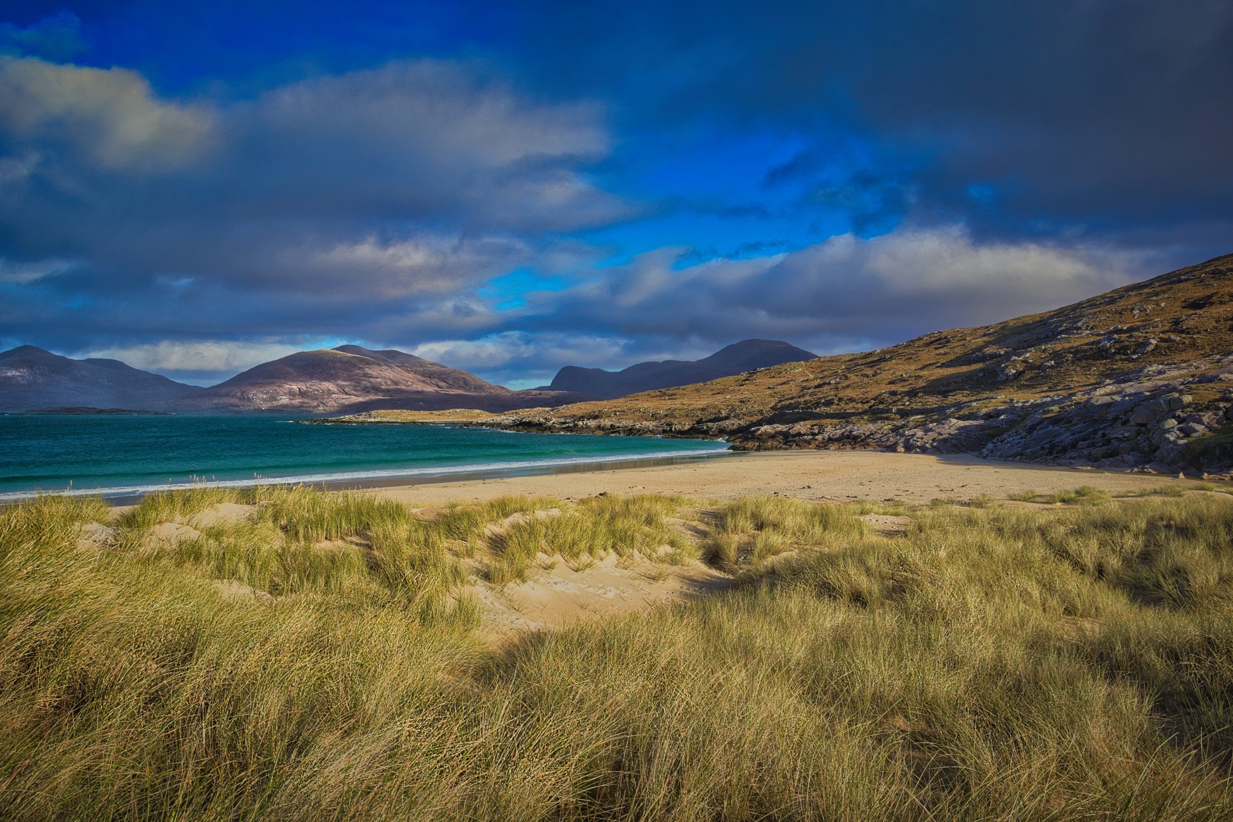 The sweeping white sands and turquoise waters of Luskentyre Beach on the Isle of Harris, framed by rugged dunes and distant mountain peaks under a dramatic sky. Fine art Scottish landscape photography capturing the vibrant coastal colours of the Oute